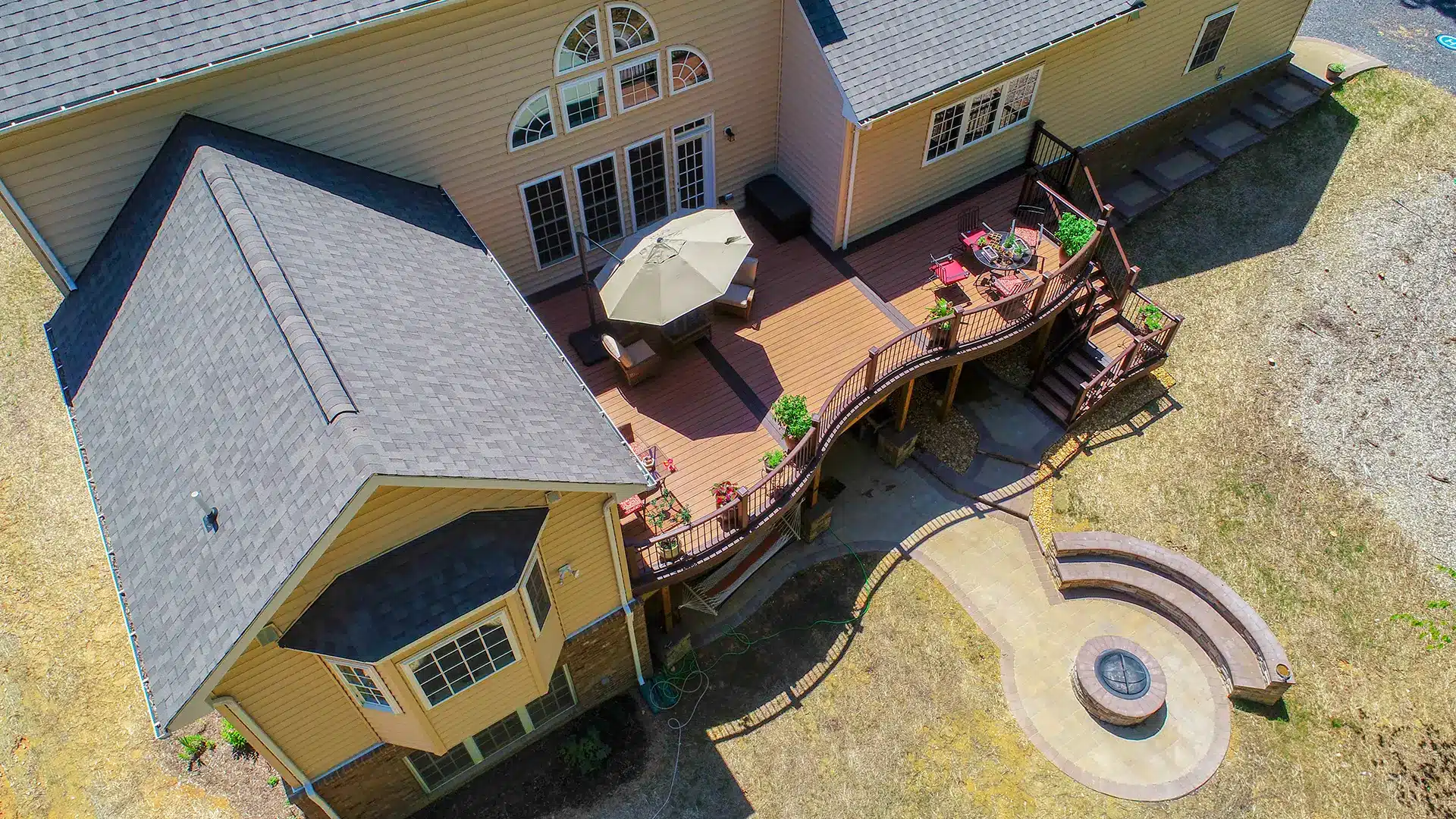 An aerial view shows a two-story tan house with a multi-level brown deck, an outdoor dining area with a patio umbrella, and a circular stone fire pit with curved seating in the backyard.