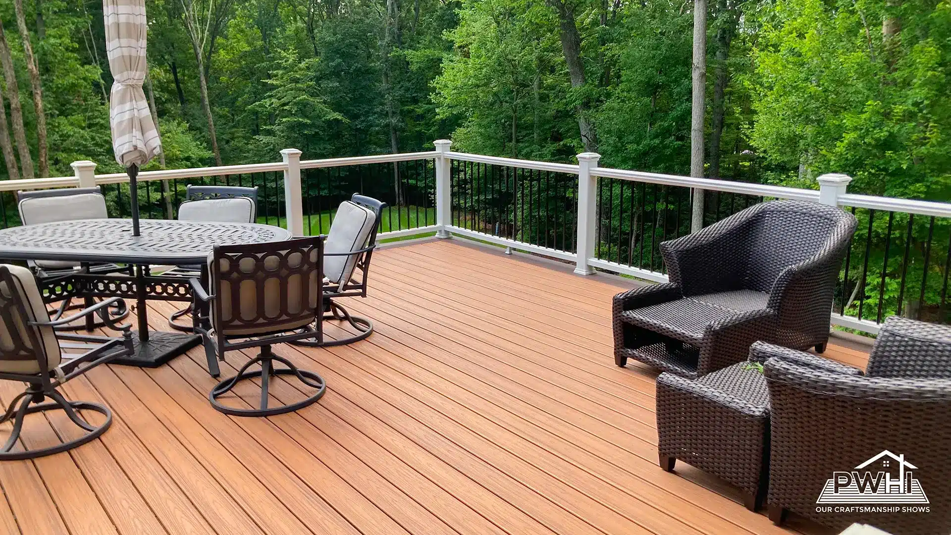 Composite deck with warm wood tone decking, white posts, black balusters, outdoor dining furniture, and wicker seating overlooking a wooded backyard.