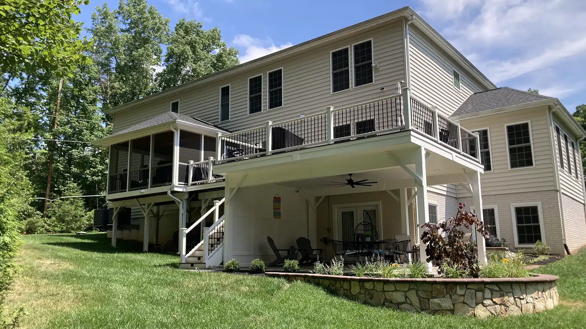 The rear of a multi-story house featuring a large elevated deck with white railings. Below the deck is a covered patio area with seating and a ceiling fan, adjacent to a stone retaining wall and landscaped garden. A screened-in porch is visible to the left of the main deck.