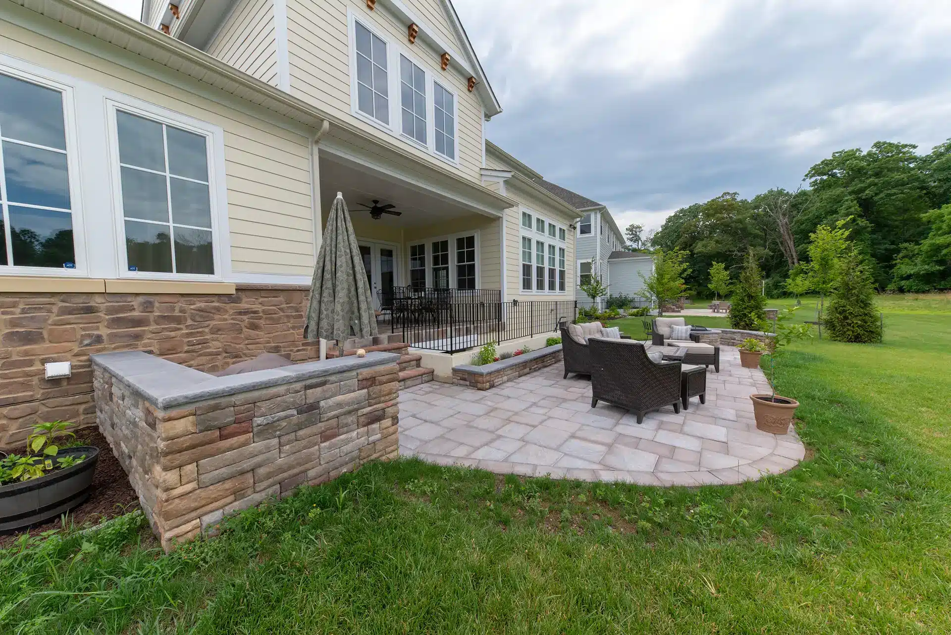 A paver patio with dark wicker outdoor furniture and a stone retaining wall attached to the back of a large, light-colored house in Fredericksburg, VA.