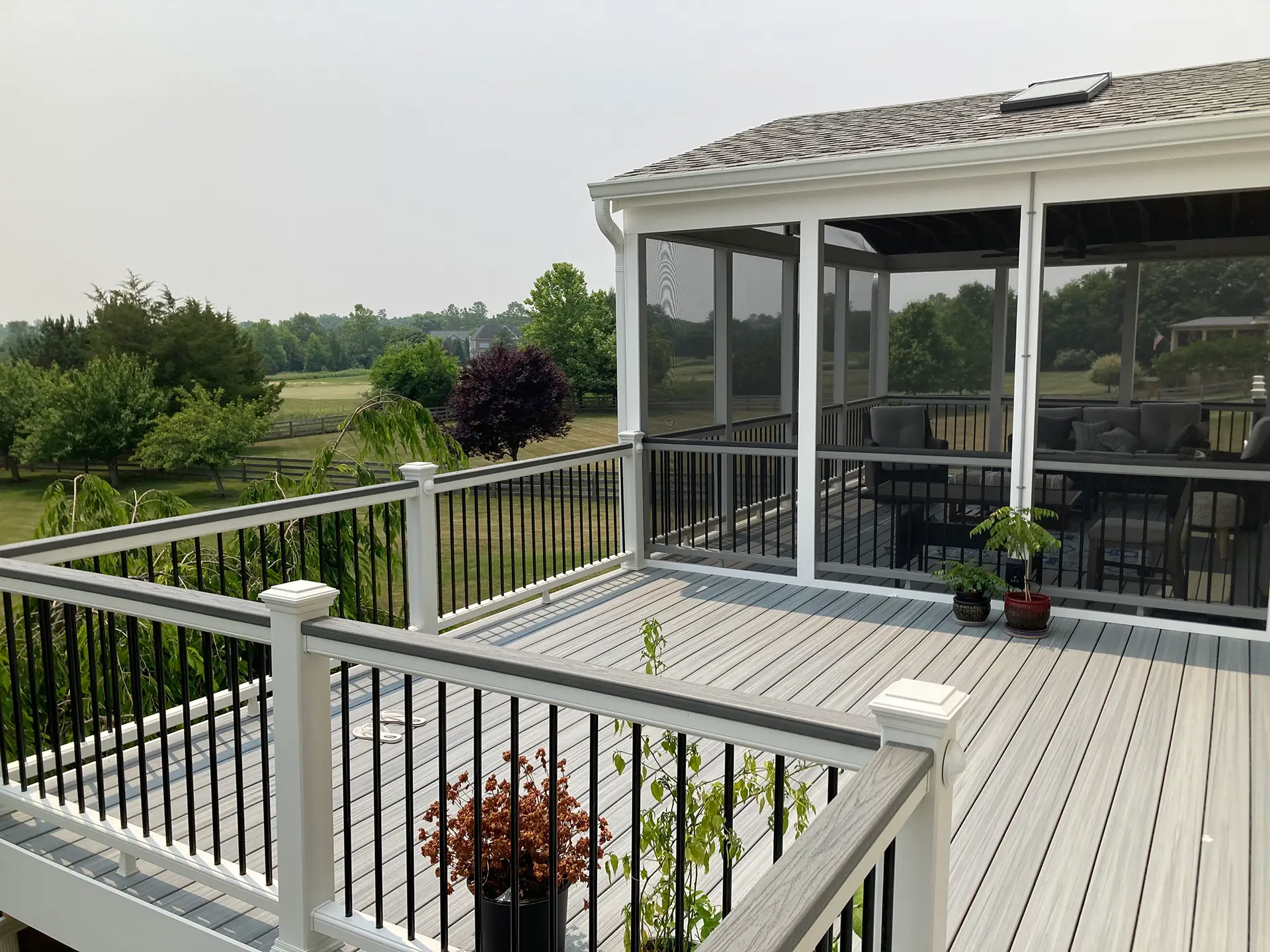 An elevated outdoor living space featuring a gray composite deck and an attached white screened-in porch with a shingled roof. The deck is enclosed by a white railing with black balusters and includes several potted plants.