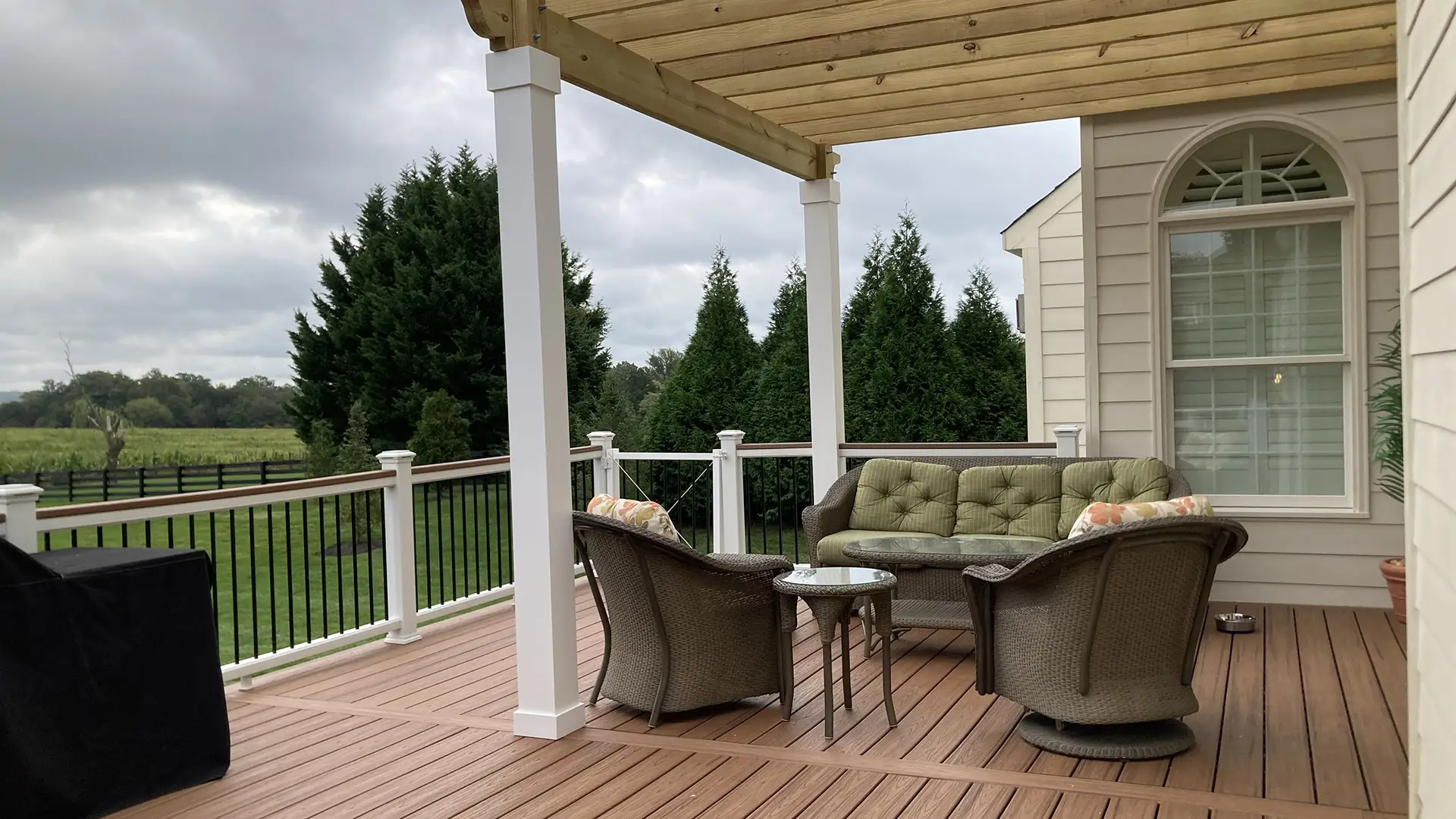 Composite deck with warm brownish tone decking, white posts, black balusters, a wood pergola, and wicker seating overlooking a green backyard.