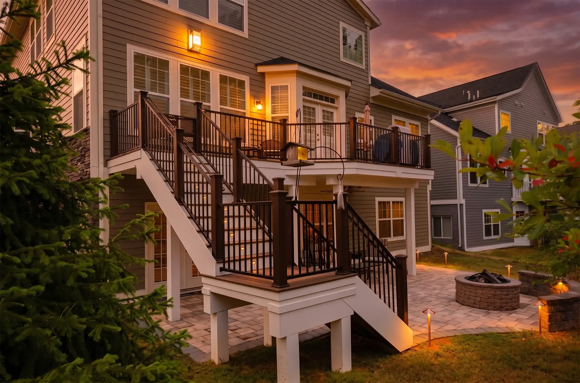 The rear exterior of a multi-story home at dusk, featuring a white elevated deck with dark railings and a matching staircase that leads down to a stone patio. The patio area includes a circular stone fire pit and several lit landscape path lights.