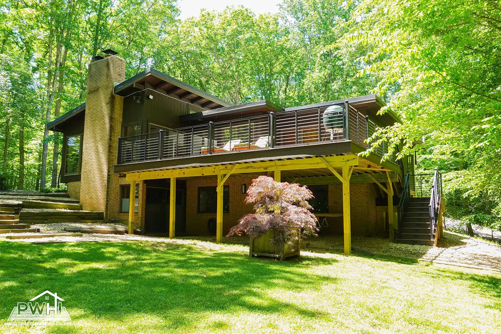 A large deck with a modern cable railing system is attached to the rear of a two-story home. The house features dark siding on the upper level, a brick lower level, and a prominent brick chimney. The structure is surrounded by lush green trees and a grassy lawn.