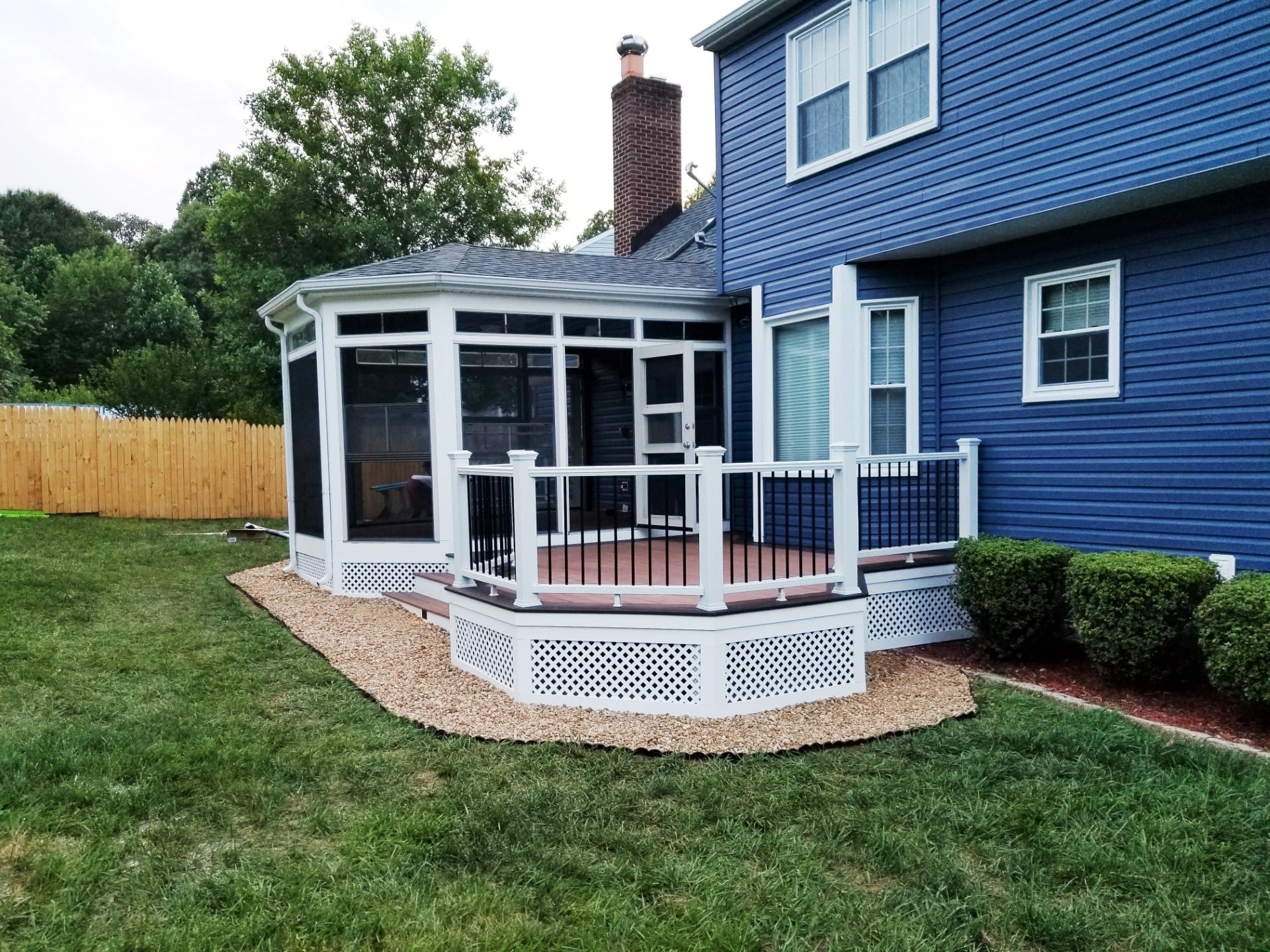 deck construction in germantown md The exterior of a blue two-story house with a white screen room addition. A multi-level deck with white railings and black balusters extends from the house, surrounded by light-colored gravel and a green lawn. White lattice covers the base of both the sunroom and the deck.