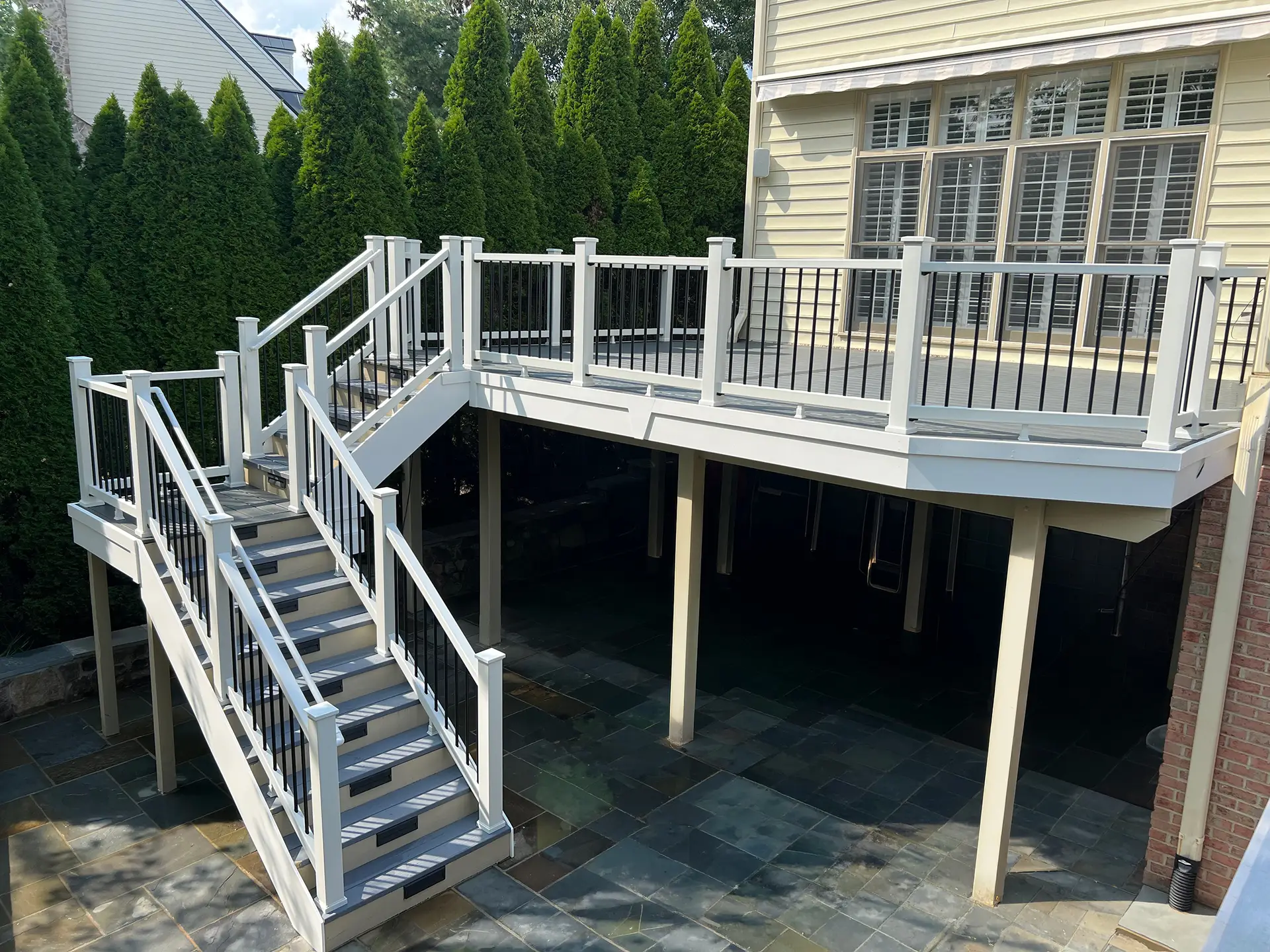 deck construction in haymarket va Raised composite deck with gray decking, white railings, black balusters, and a wide staircase leading down to a stone patio.