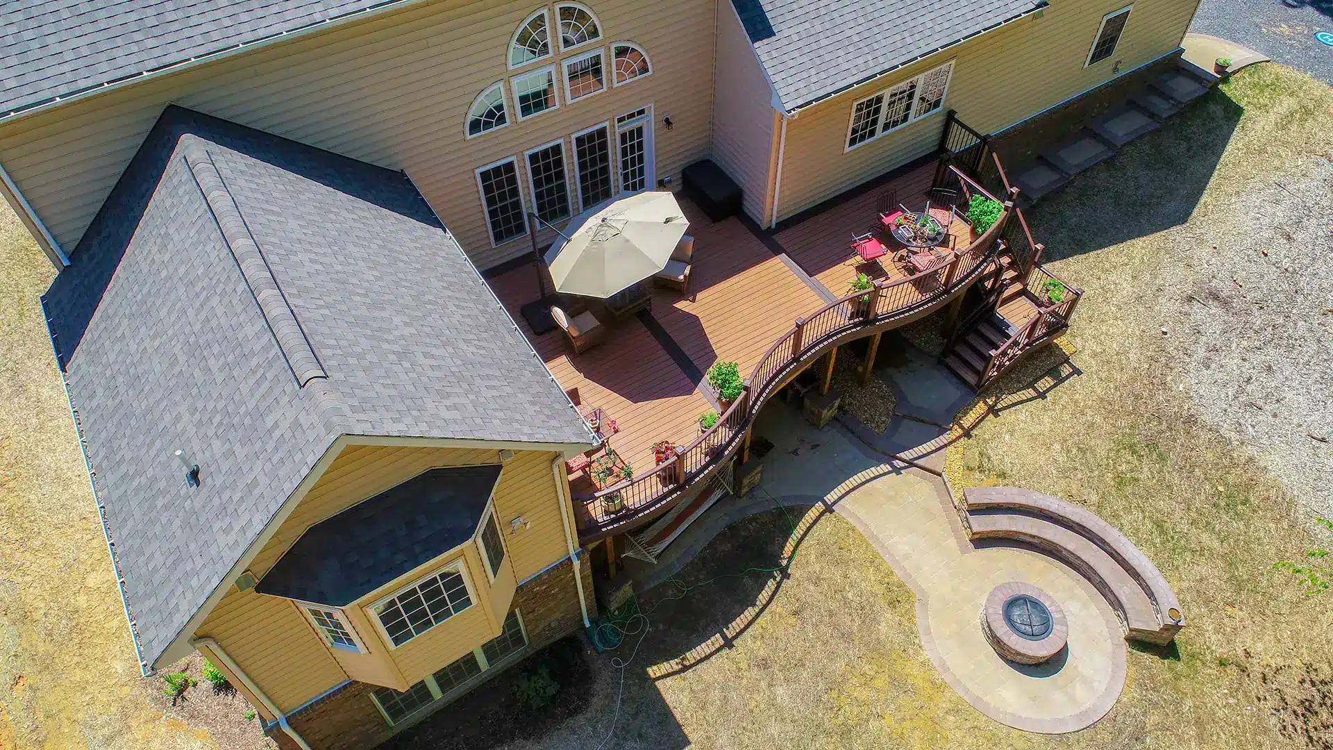 deck construction in lorton va An aerial view shows a two-story tan house with a multi-level brown deck, an outdoor dining area with a patio umbrella, and a circular stone fire pit with curved seating in the backyard.