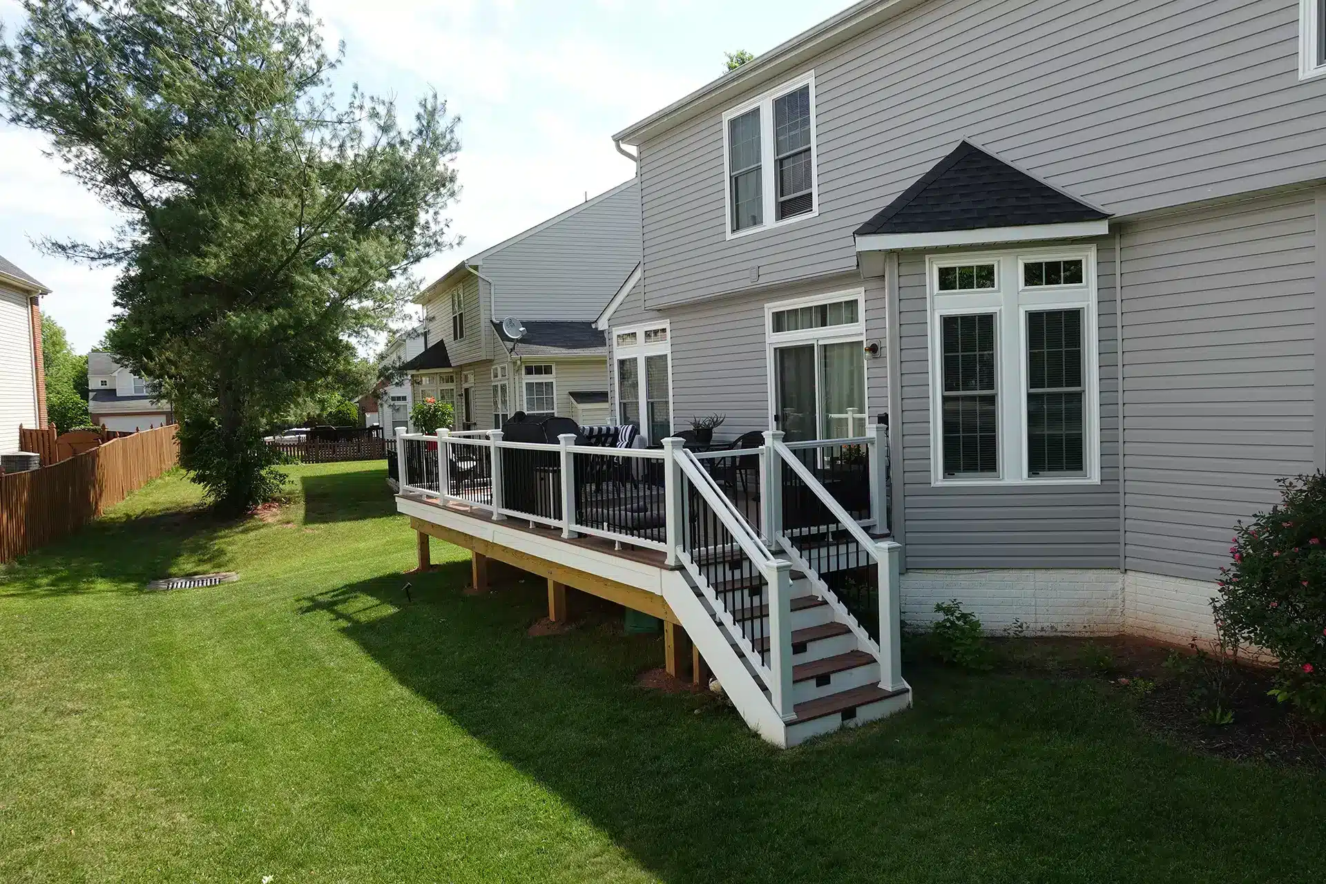 deck construction in woodbridge va Raised composite deck with brown decking, white railings, black balusters, outdoor seating, and stairs leading down to a grassy backyard.