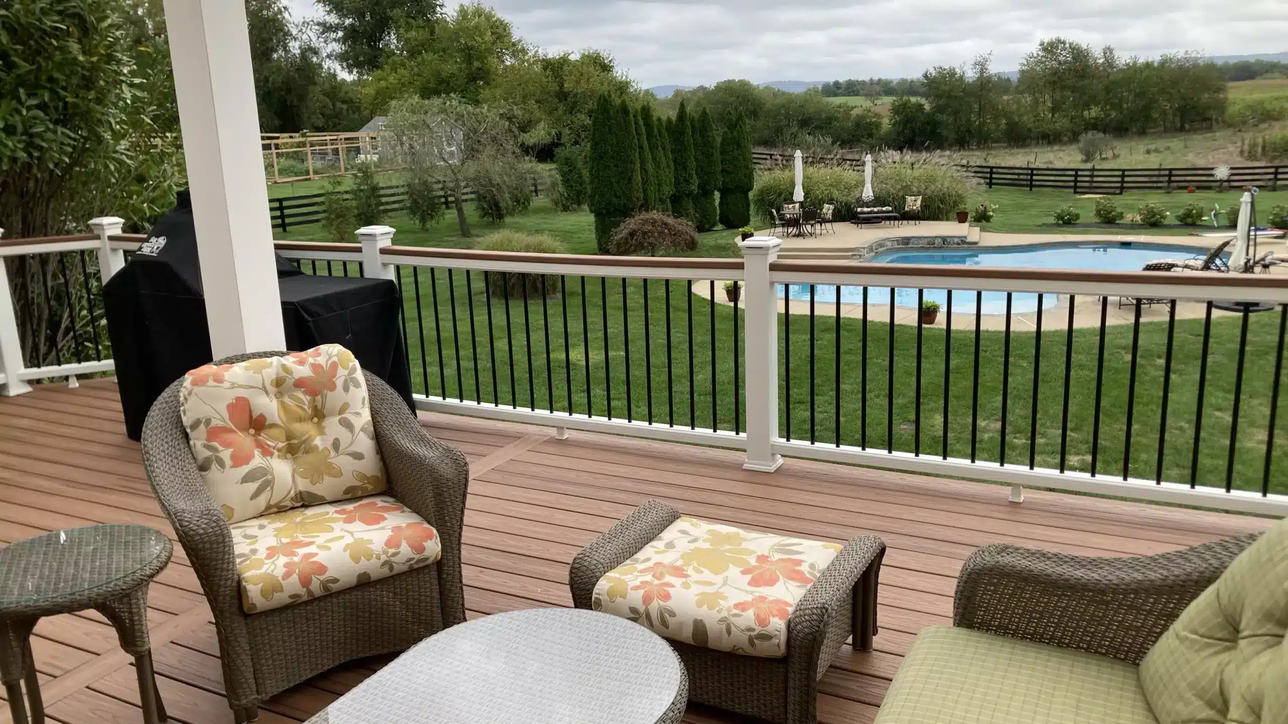deck contractor in alexandria va An elevated outdoor deck featuring composite decking in a brown tone. The deck is furnished with woven wicker-style seating, including an armchair and ottoman with orange and yellow floral cushions, alongside a small side table and a larger coffee table