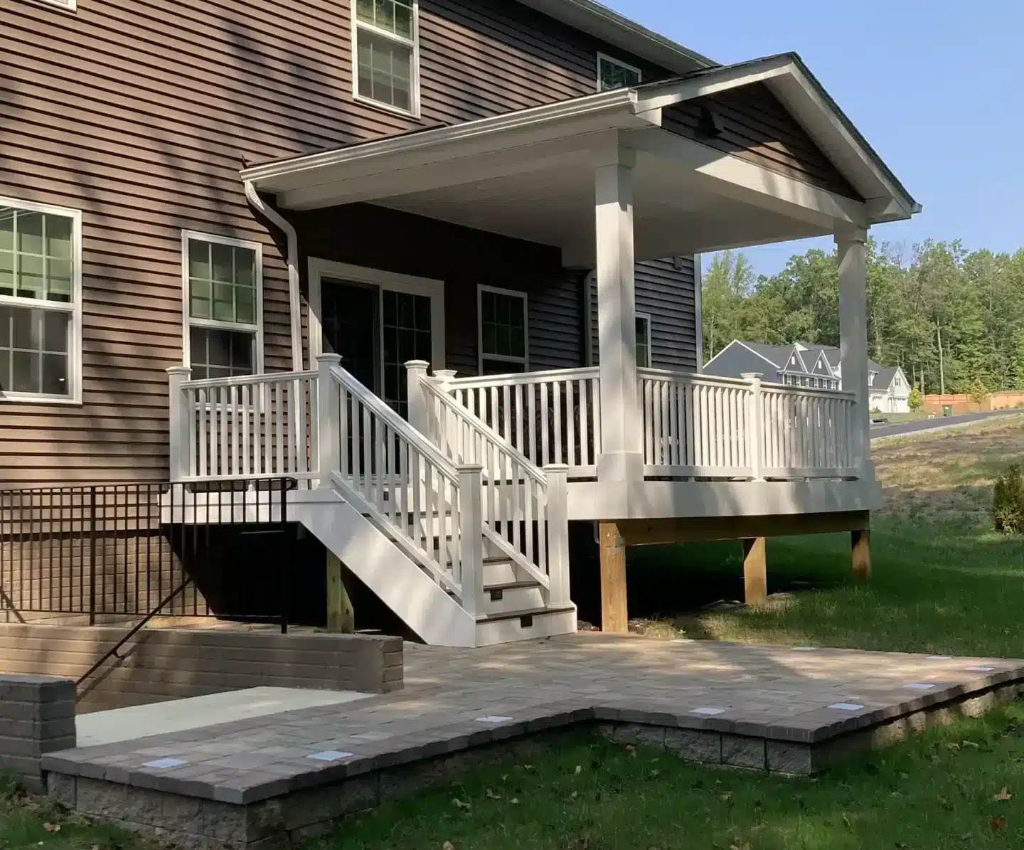 deck contractor in bowie md Covered raised deck with white railings, stairs, and a gable roof overlooking a paved patio and grassy backyard.