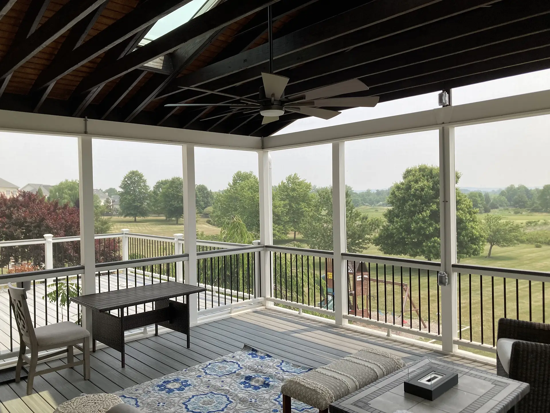 A screened-in porch with dark rafters and a ceiling fan overlooks a lush green yard. The porch features grey composite decking, a patterned outdoor rug, and various pieces of furniture, including a table and seating.