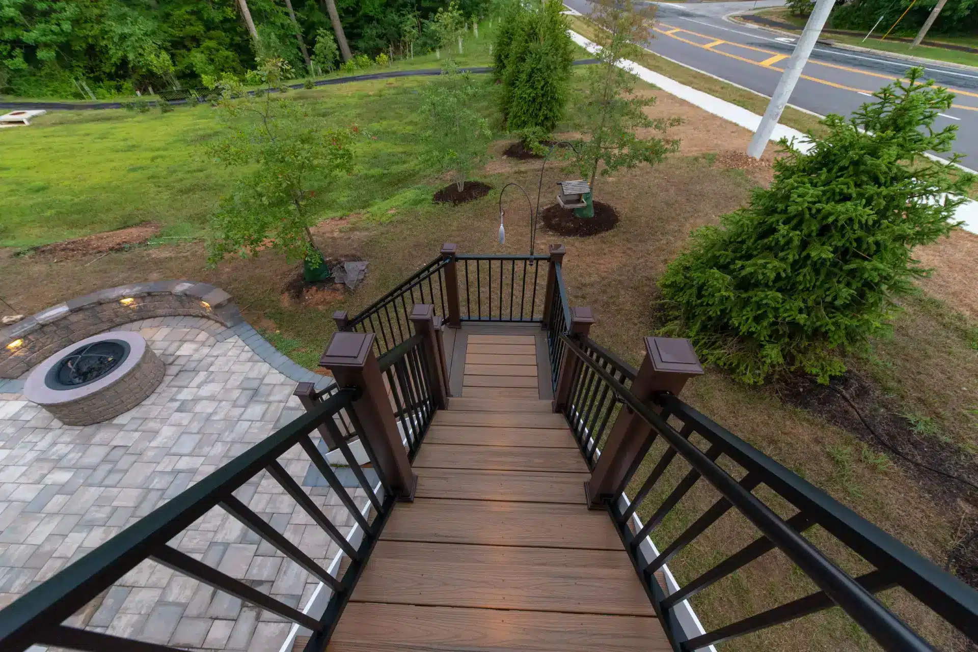 deck contractor in gaithersburg md A high-angle, downward view of a brown composite deck staircase leading to a backyard. The stairs are flanked by black metal railings with dark brown posts. To the left of the stairs is a circular stone fire pit built into the paved patio.
