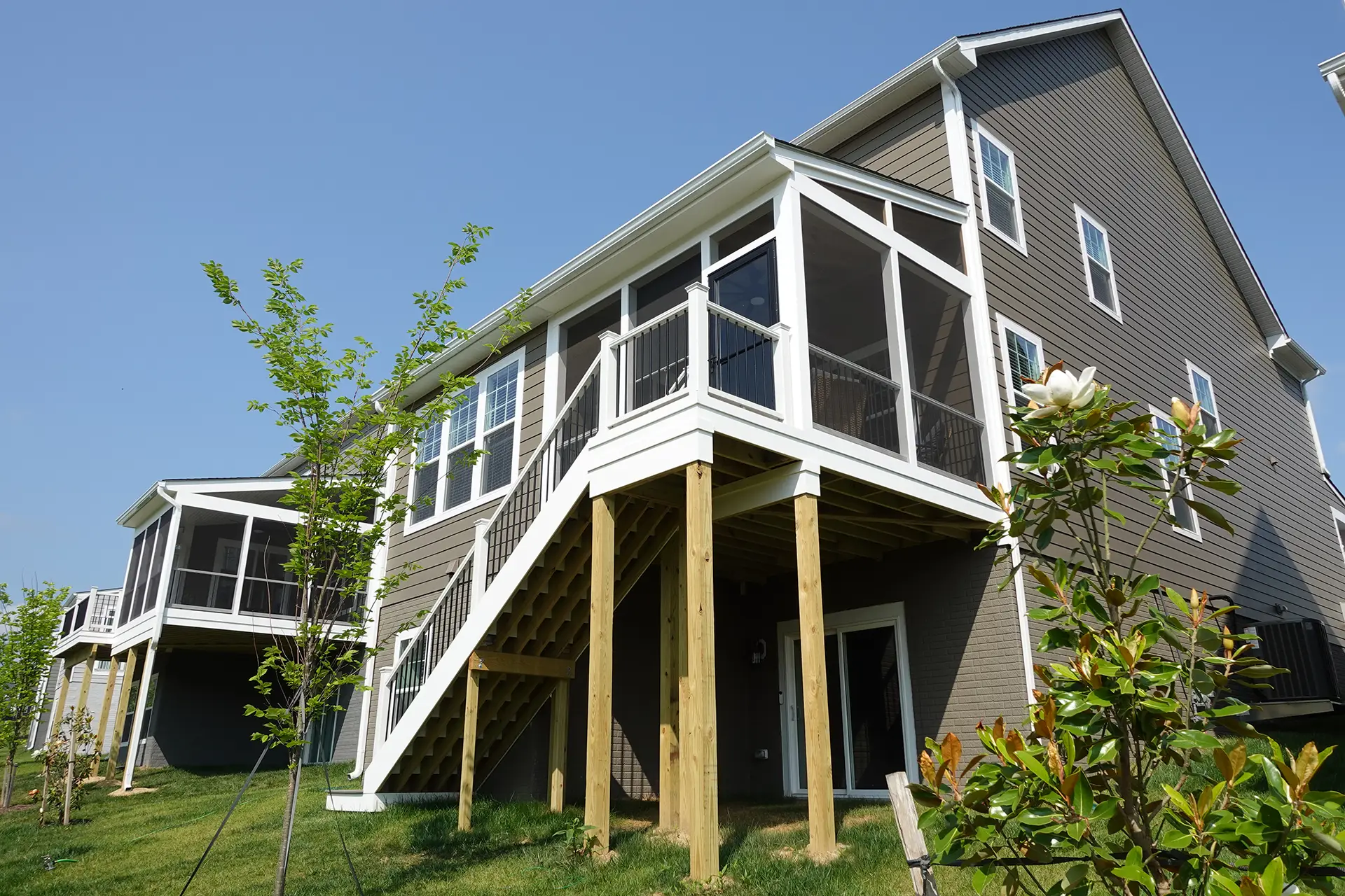 deck contractor in manassas va The rear exterior of a multi-story residential building in Manassas, VA, with grey siding and white trim. The focus is on a raised, white screened-in porch supported by tall posts. A set of stairs with white railings and dark balusters leads from the ground level up to the porch.