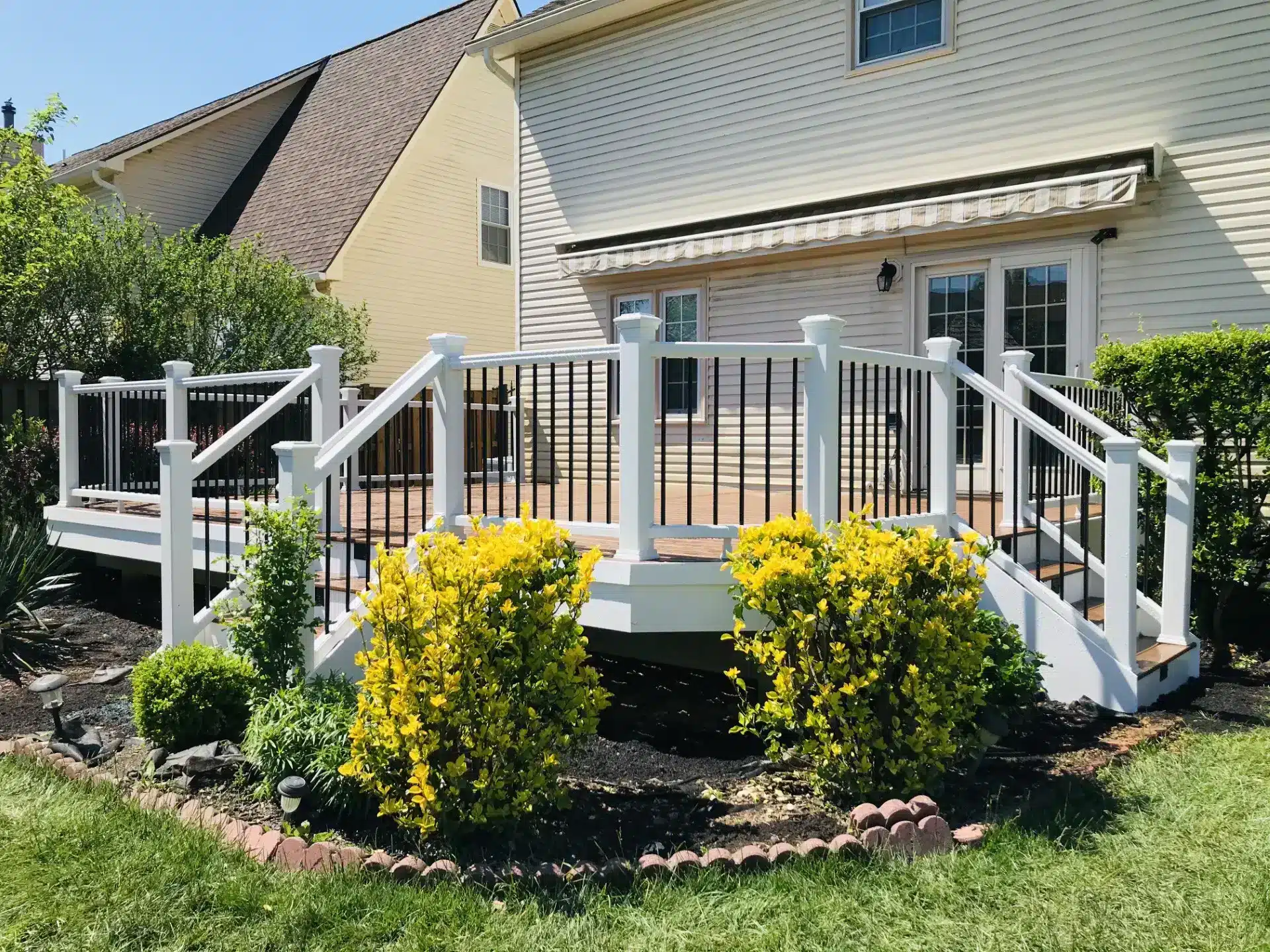 deck contractor in reston va A black-and-white railing surrounds a composite deck attached to the back of a light-colored house in Reston, VA. Bright yellow flowering bushes and other green plants sit in a mulched garden bed in the foreground.