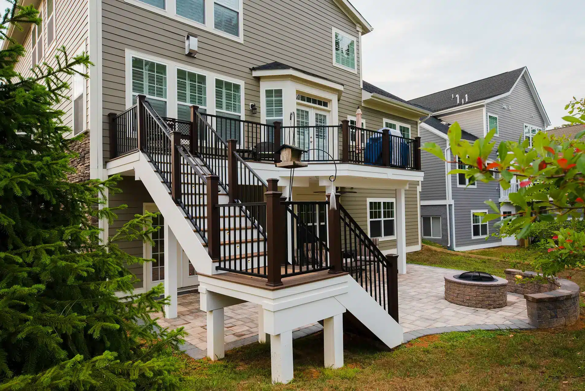 deck contractor in woodbridge va An elevated composite deck with black railings and a white staircase leading down to a stone paver patio featuring a built-in fire pit in Woodbridge, VA.