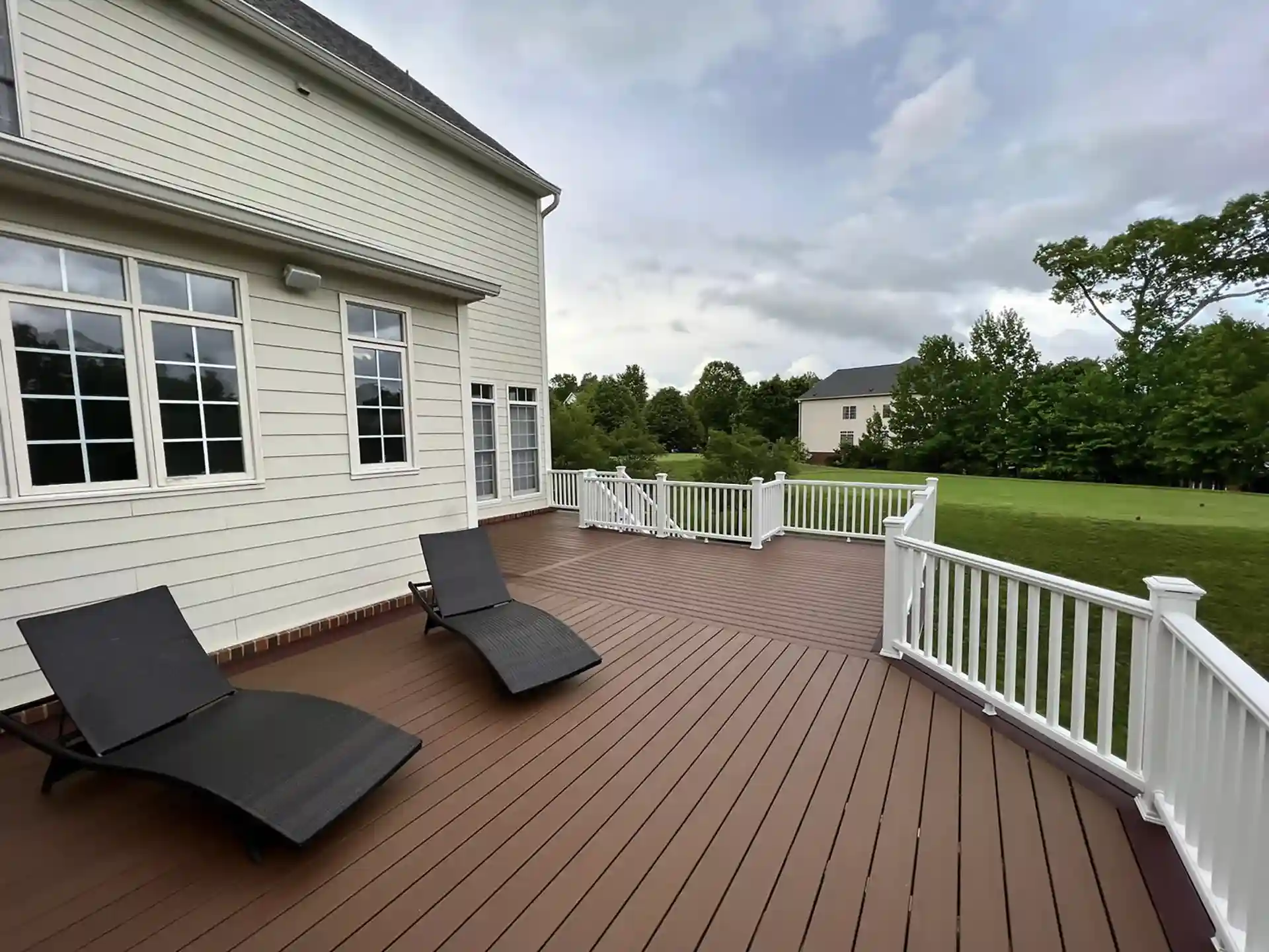 Spacious brown composite deck with white railings featuring black lounge chairs and overlooking a lush green backyard.
