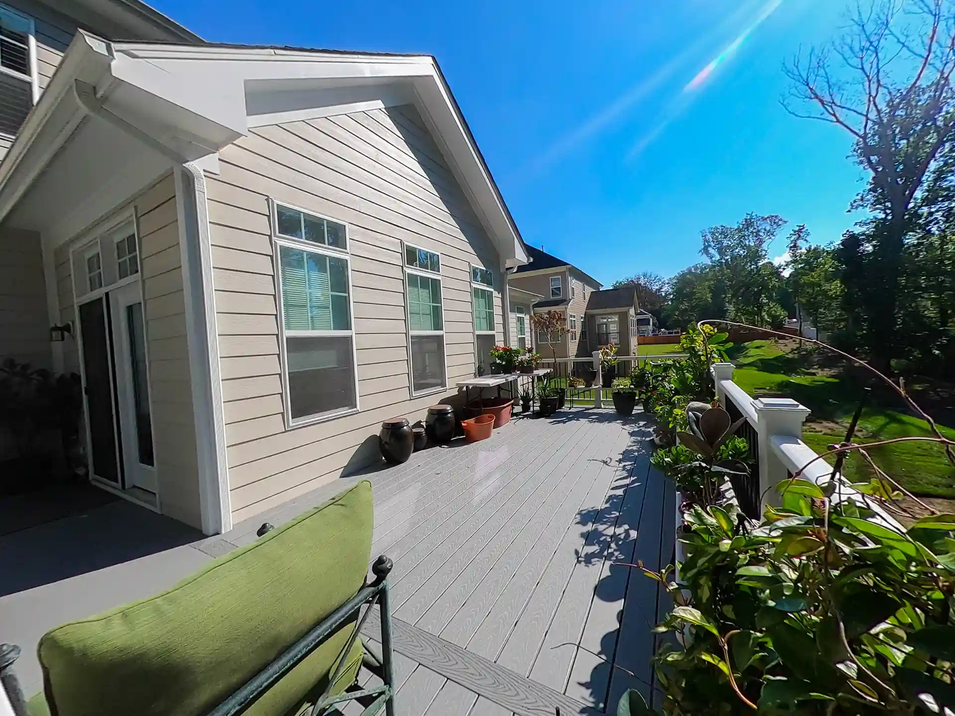 Sunny backyard deck with potted plants, outdoor seating, and white railings attached to a beige house with large windows.
