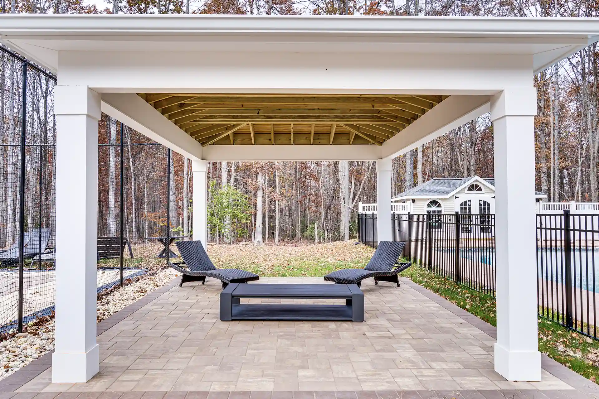 Stylish covered patio with a paver floor, lounge chairs, and a modern fire pit overlooking a wooded backyard.
