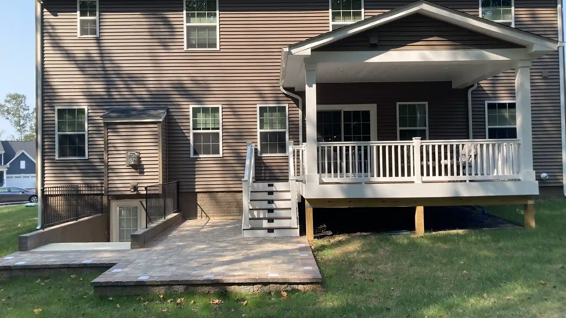 Backyard view of a house featuring a raised covered deck with white railings, stairs leading down to a paver patio.
