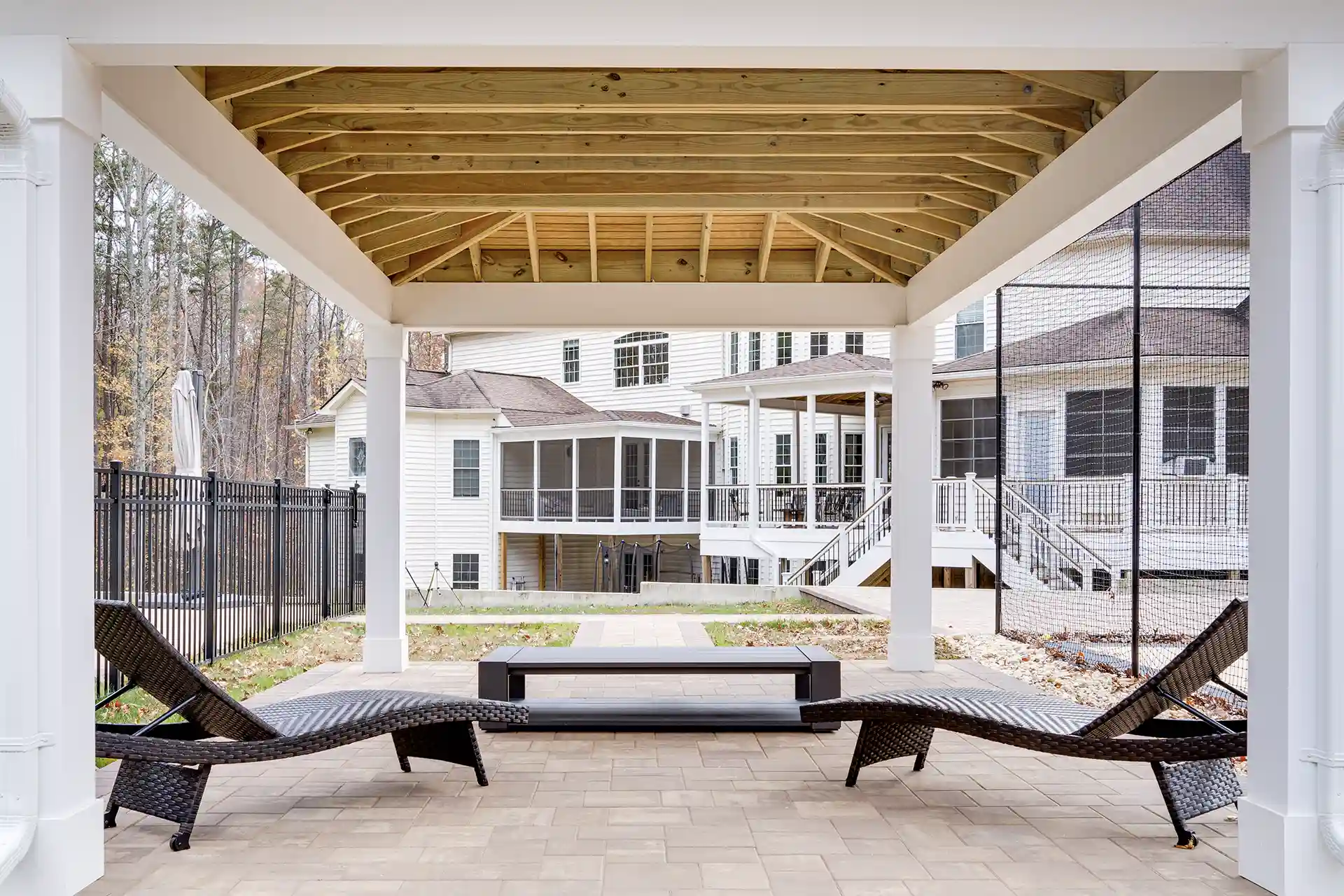 Covered patio with a wooden ceiling, supported by white columns, and two wicker lounge chairs on a paved floor. 
