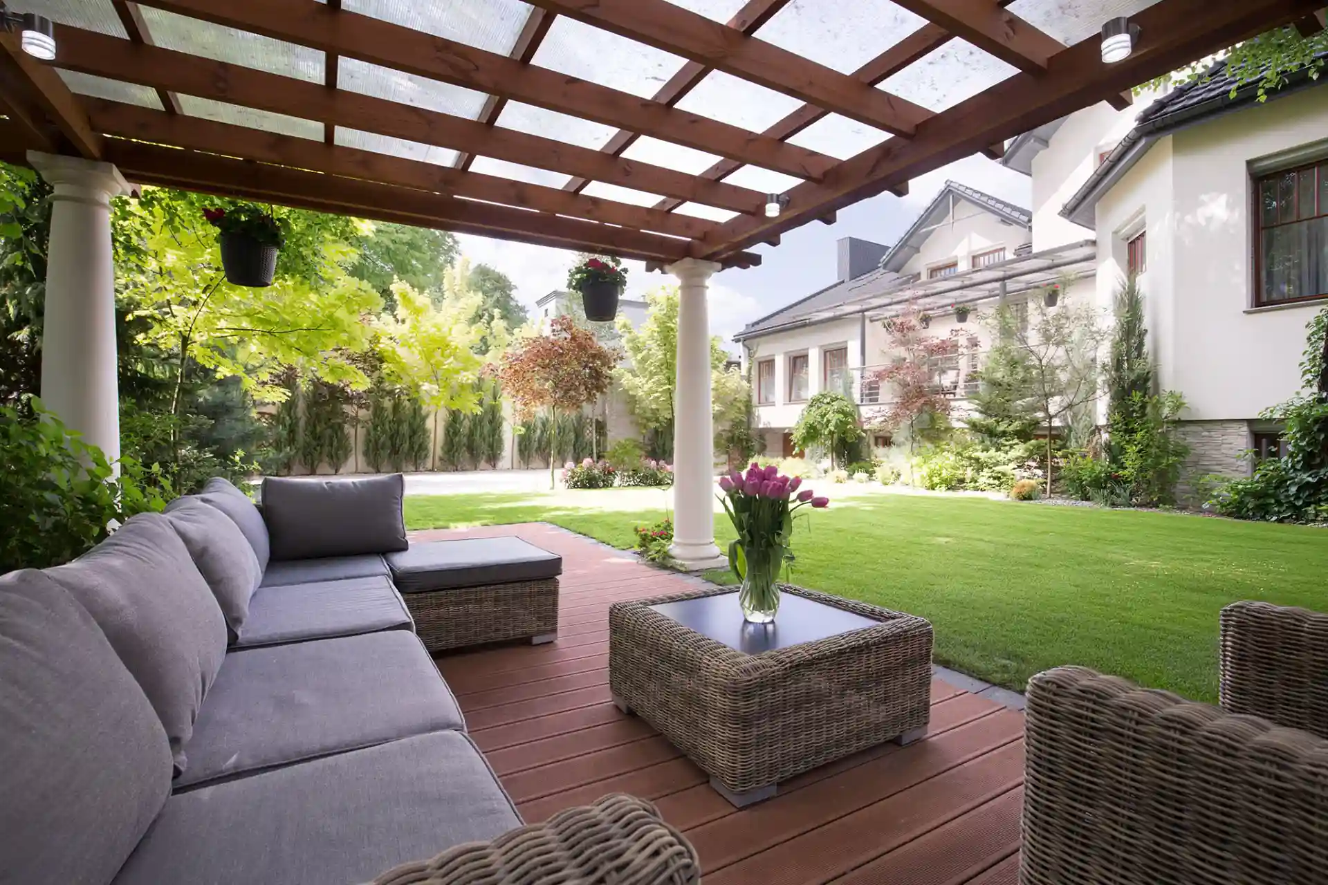 Shaded patio with a wooden pergola, gray seating, and lush greenery. 