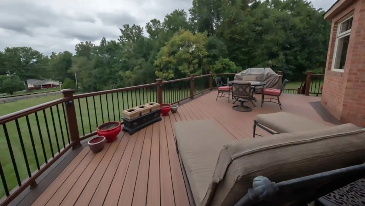 Composite deck with railing, outdoor furniture, and planters, adjacent to a brick house overlooking a grassy backyard with trees.