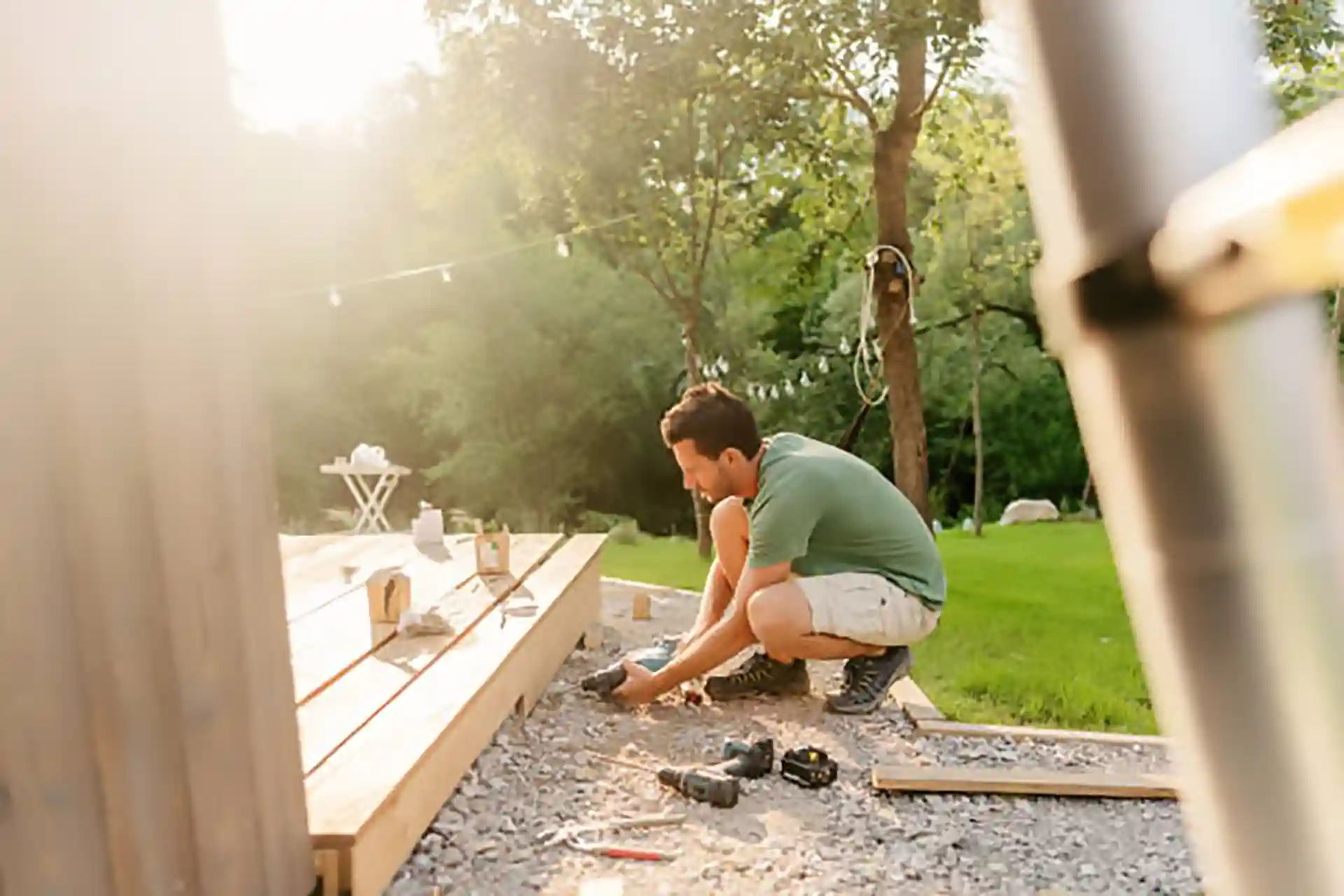 Person installing deck framing on gravel base using tools in a backyard with trees and string lights 