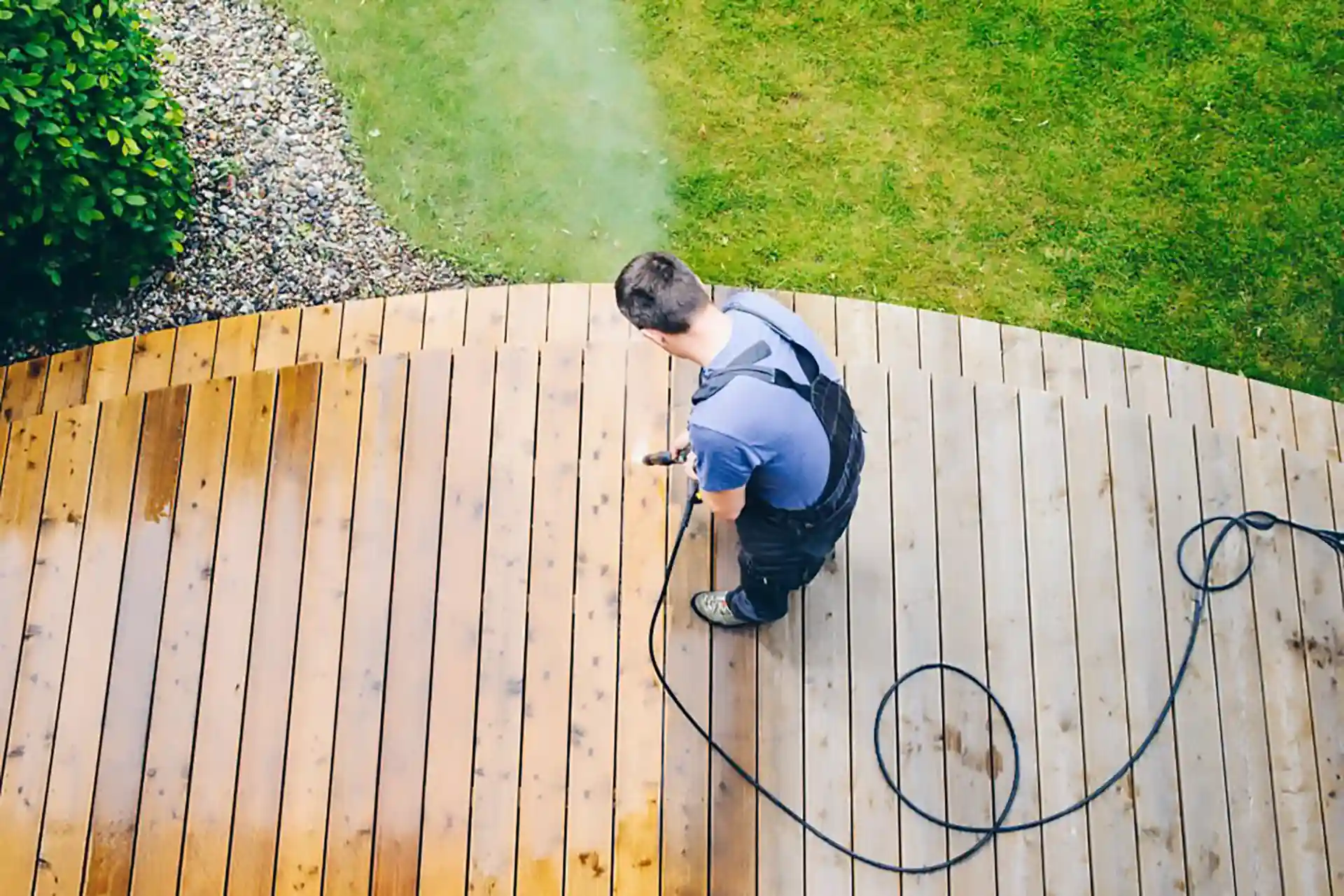 Person using a pressure washer to clean a curved wooden deck next to grass and gravel landscaping 