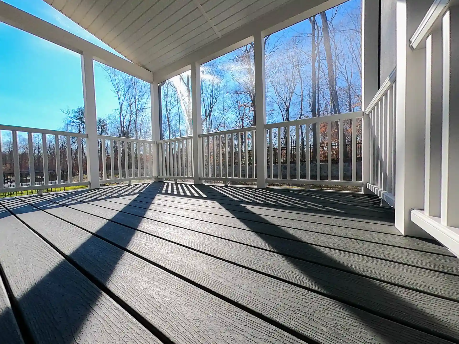 Covered porch with gray composite decking, white railing, and sunlight casting shadows on the floor, surrounded by bare trees. 