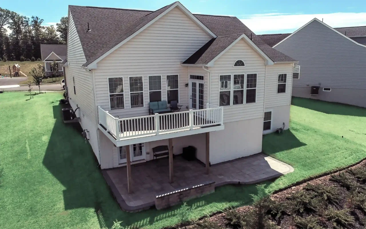 Elevated white-railed deck attached to a beige two-story house with a concrete patio underneath and surrounding grass lawn.