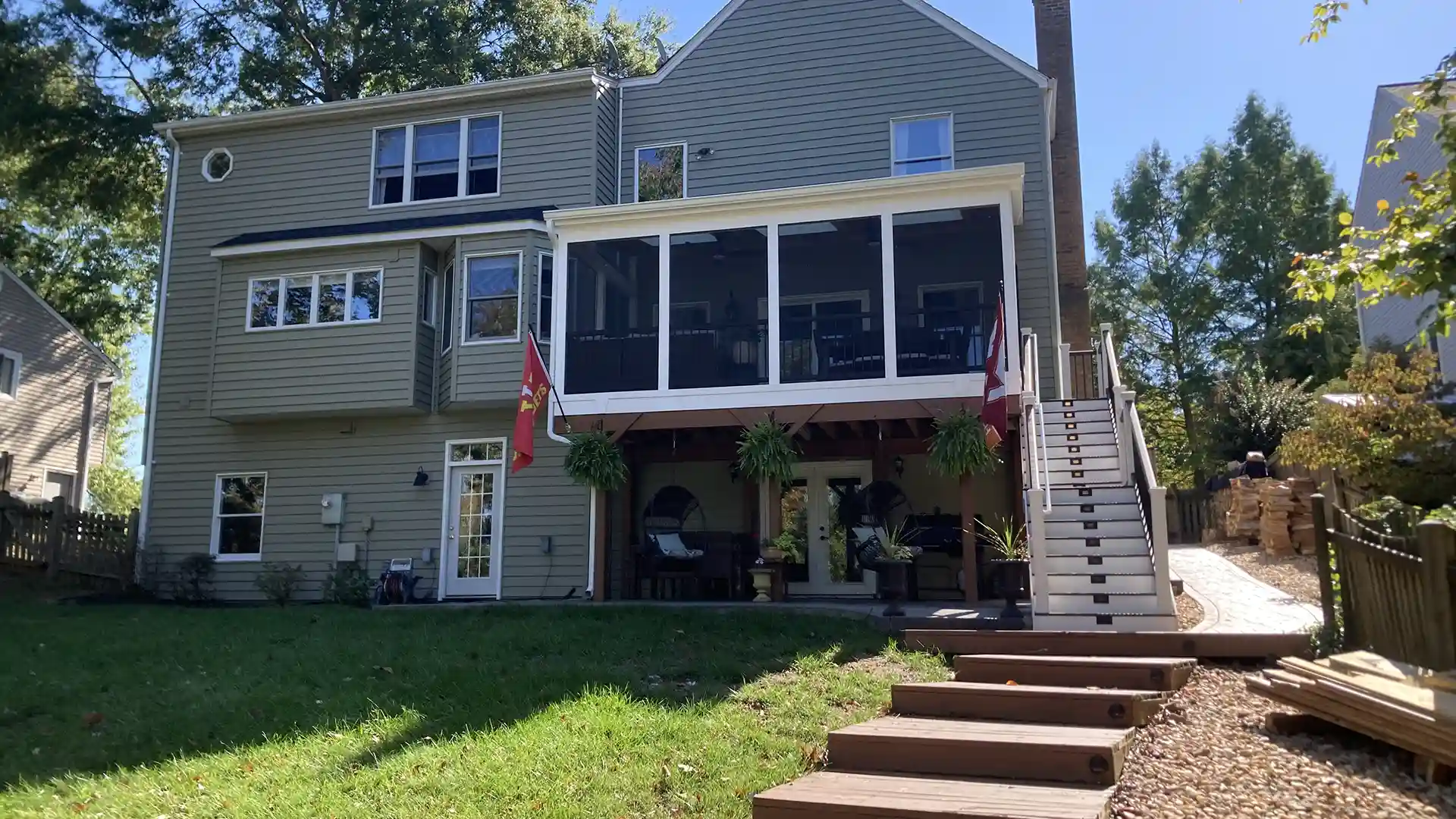 Screened-in upper deck with exterior staircase attached to a three-story house, with seating area and potted plants beneath.
