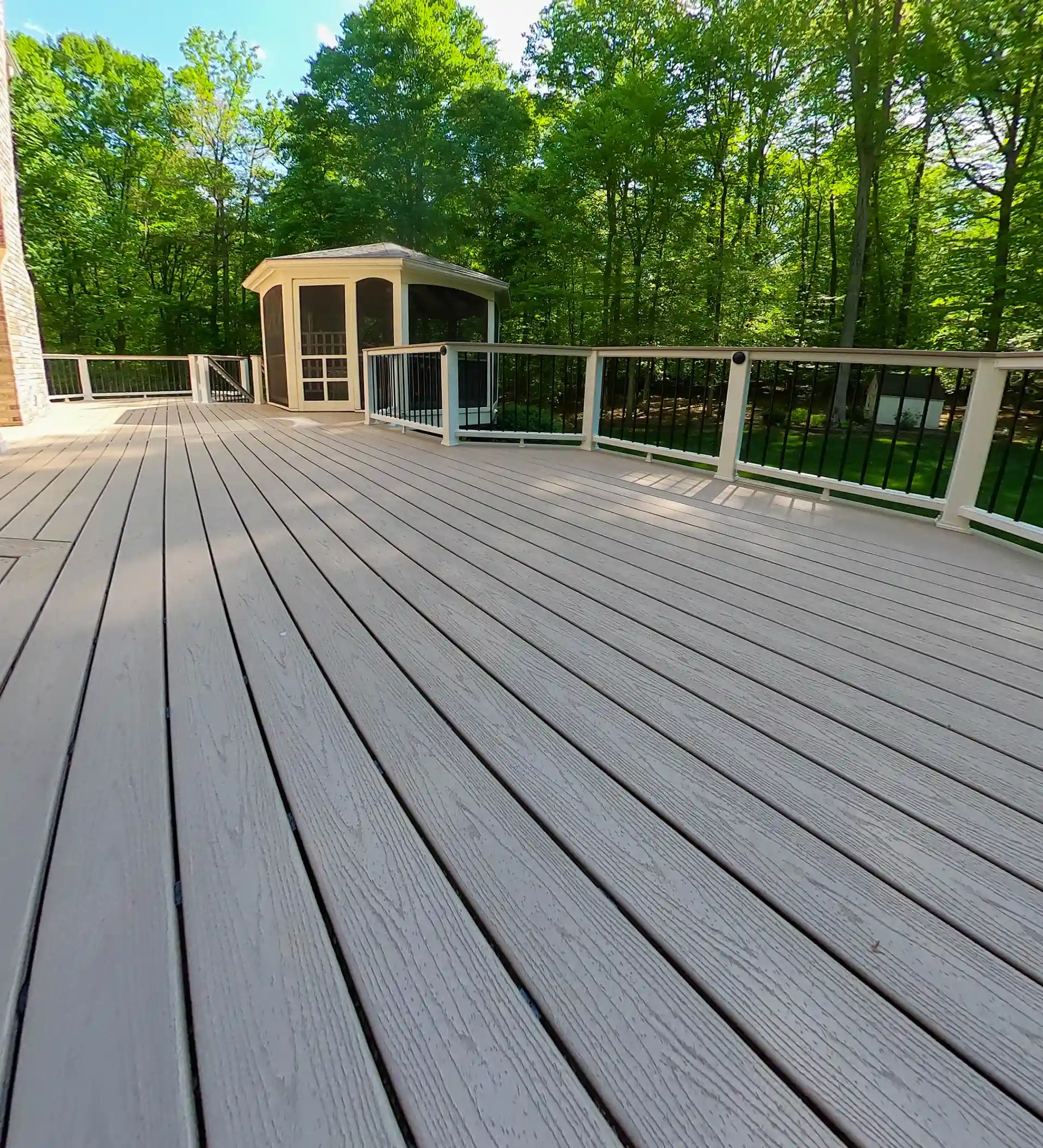 Light-colored composite decking with a white railing and a small gazebo in the background surrounded by trees.
