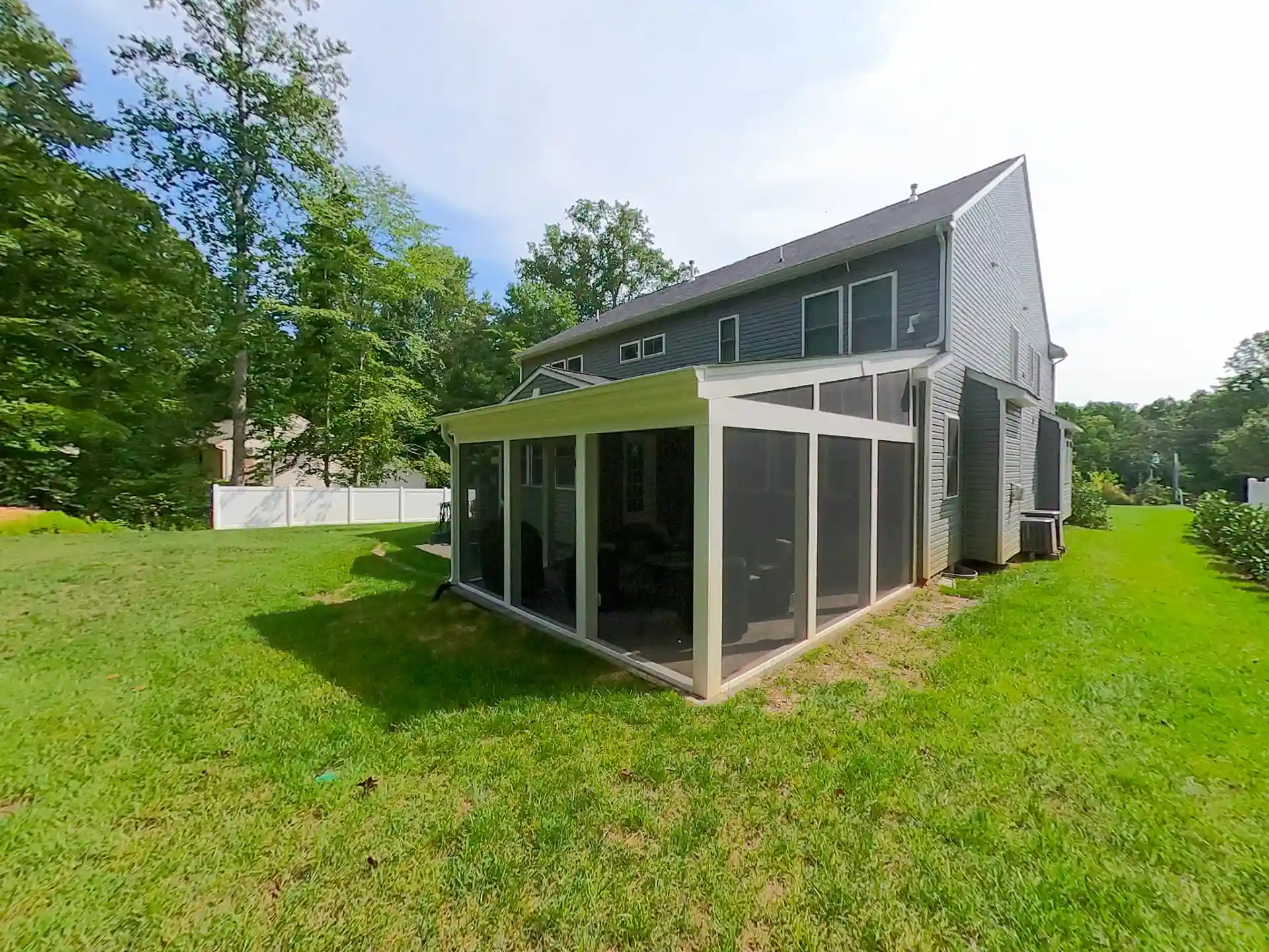 Screened porch addition attached to the back of a gray two-story house, featuring white framing and mesh screens, surrounded by a green lawn.
