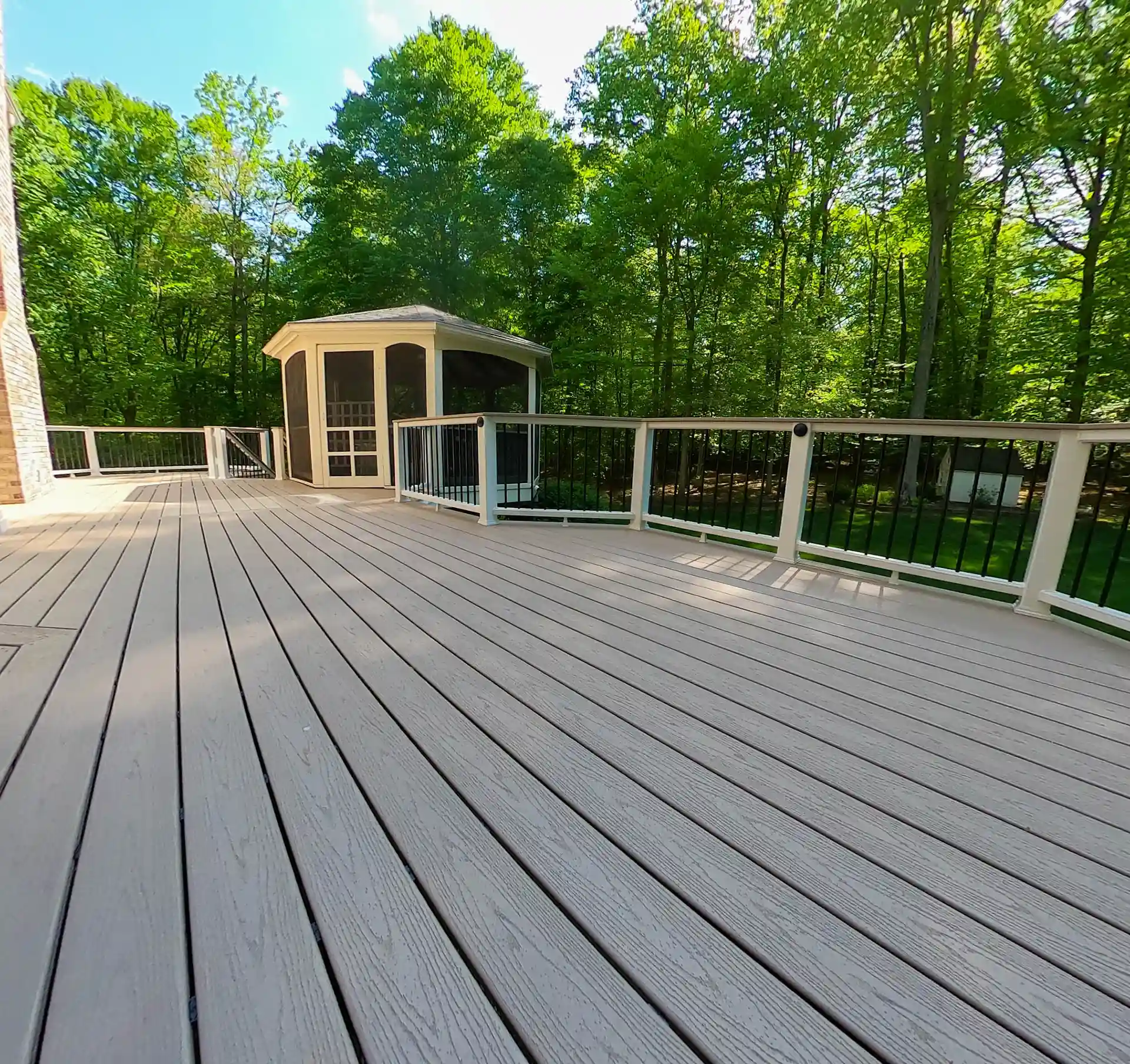 Deck with composite flooring and railing, featuring a screened-in structure in the background surrounded by trees. 