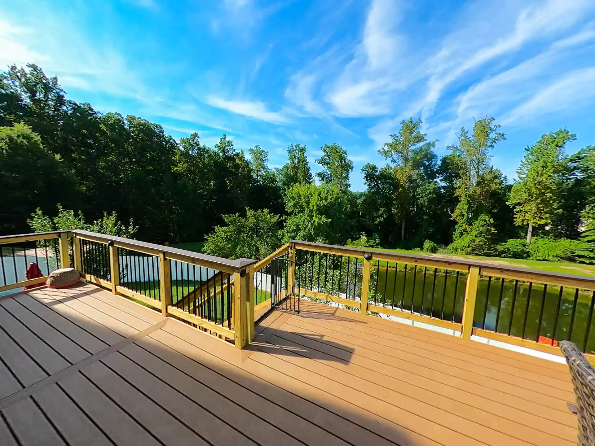 Composite deck with wood railings and black metal balusters overlooking a pond and dense green trees under a bright blue sky.
