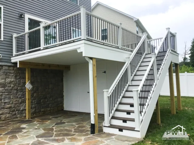 A raised deck with white and black railing, stone flooring underneath, and stairs leading to the ground. 