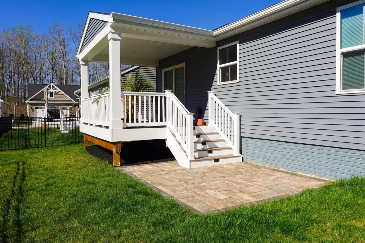 View of a white-railed deck with stairs leading down to a paved patio area, attached to a gray house with a green lawn. 