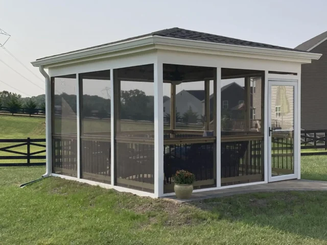Screened-In Decks vs Screen Rooms A screen room with a white frame and brown screening sits on a lush green lawn, surrounded by a black fence and rolling green hills