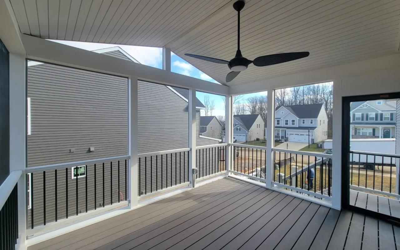 Backyard view of a house with a screened porch and a raised deck. The area below the deck has seating, and there is a stone garden wall along the grass.