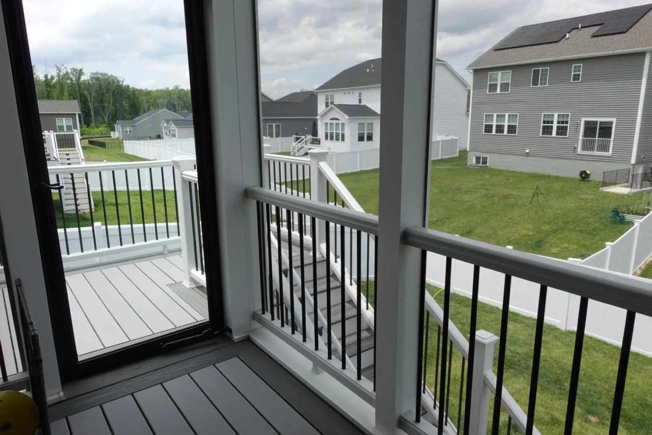 View from inside a screened-in porch with a staircase leading down to the yard. The porch has a deck with horizontal railing and a clear view of the grassy area outside.