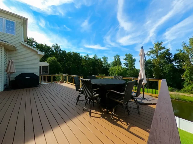 A composite outdoor deck with a dining area. The deck has light brown flooring and dark brown railings. There's a round table with chairs covered by a dark cloth in the center of the deck, and an umbrella stands nearby.