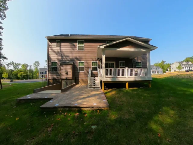 Best Deck Financing Options in Virginia A two-story, brown-sided house with a covered deck featuring white railings and stairs leading to the yard. Below the deck, there is a paved patio area with built-in lights and a small retaining wall.