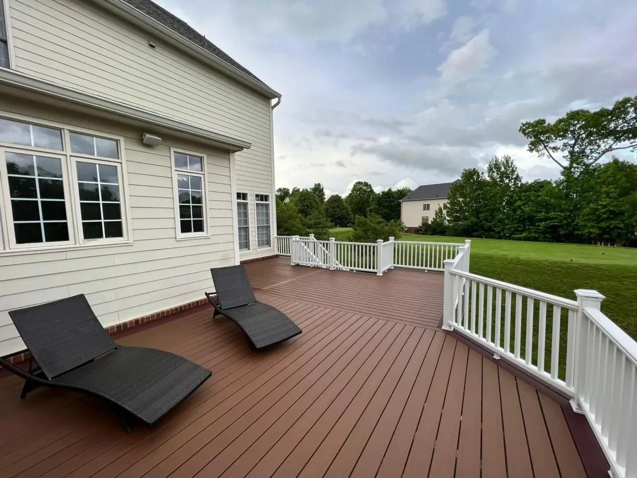 A composite deck with brown planks and white railings, featuring two dark brown lounge chairs and a matching small table.
