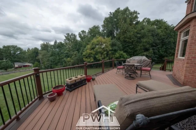 A low-angle view of a brown-sided house with a large deck and a light-colored wood pergola with string lights. The deck has black railings and is furnished with a blue rocking chair and a set of brown wicker furniture. 