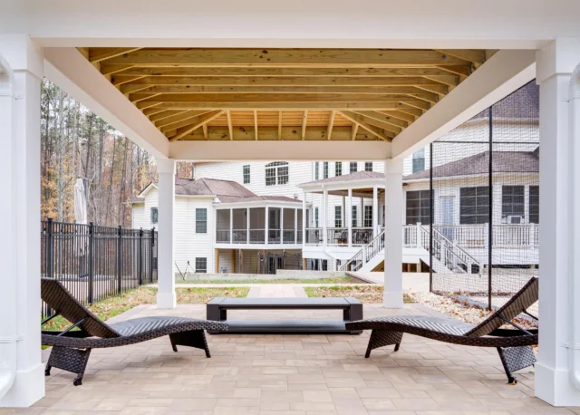 A patio featuring two dark lounge chairs and a matching low table under a white-columned pergola with a natural wood ceiling, looking out towards a large white house.