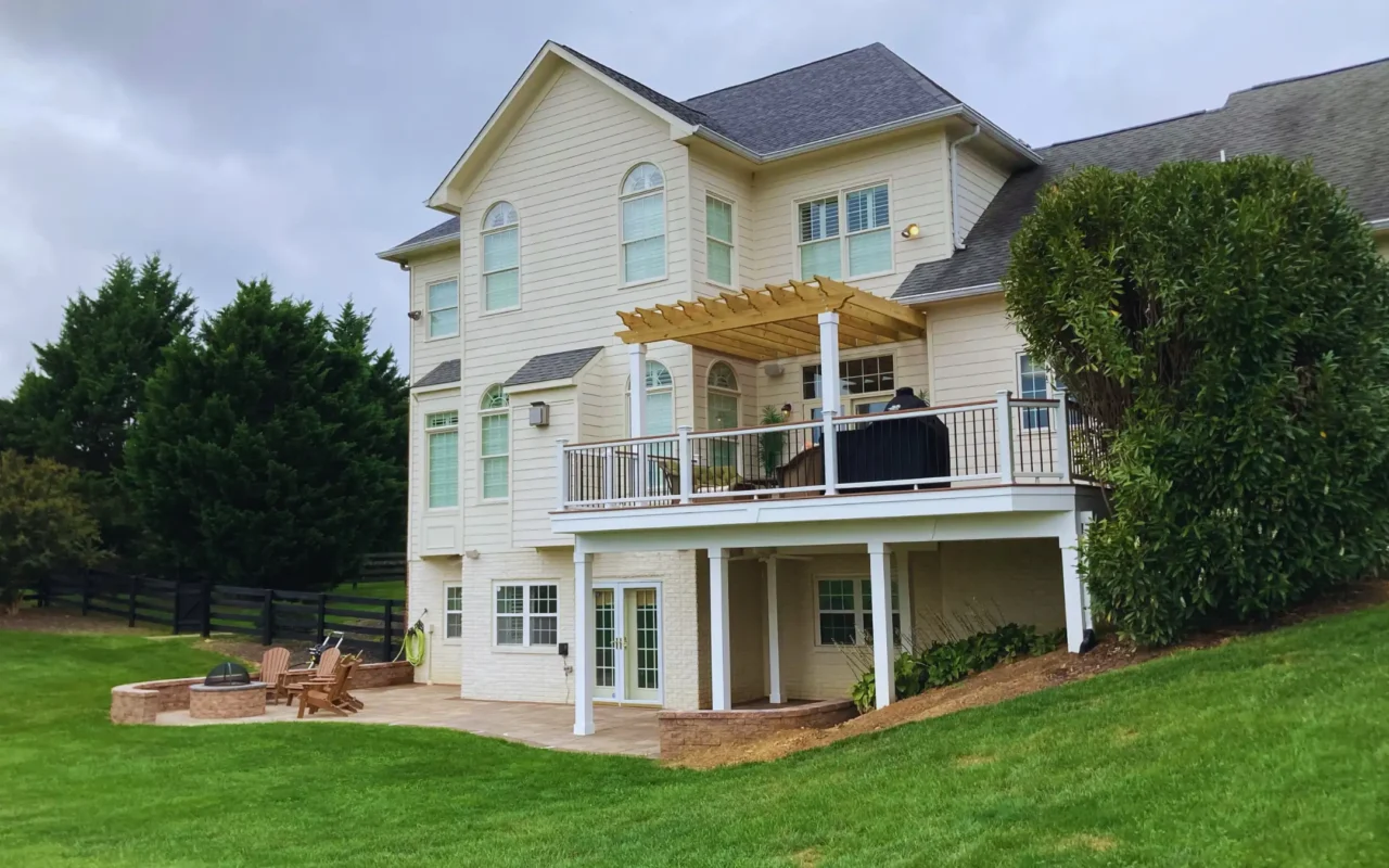Exterior of a large, light-colored house with a multi-level outdoor living space. There is a deck with a railing and a pergola, as well as a patio below with a fire pit and outdoor seating