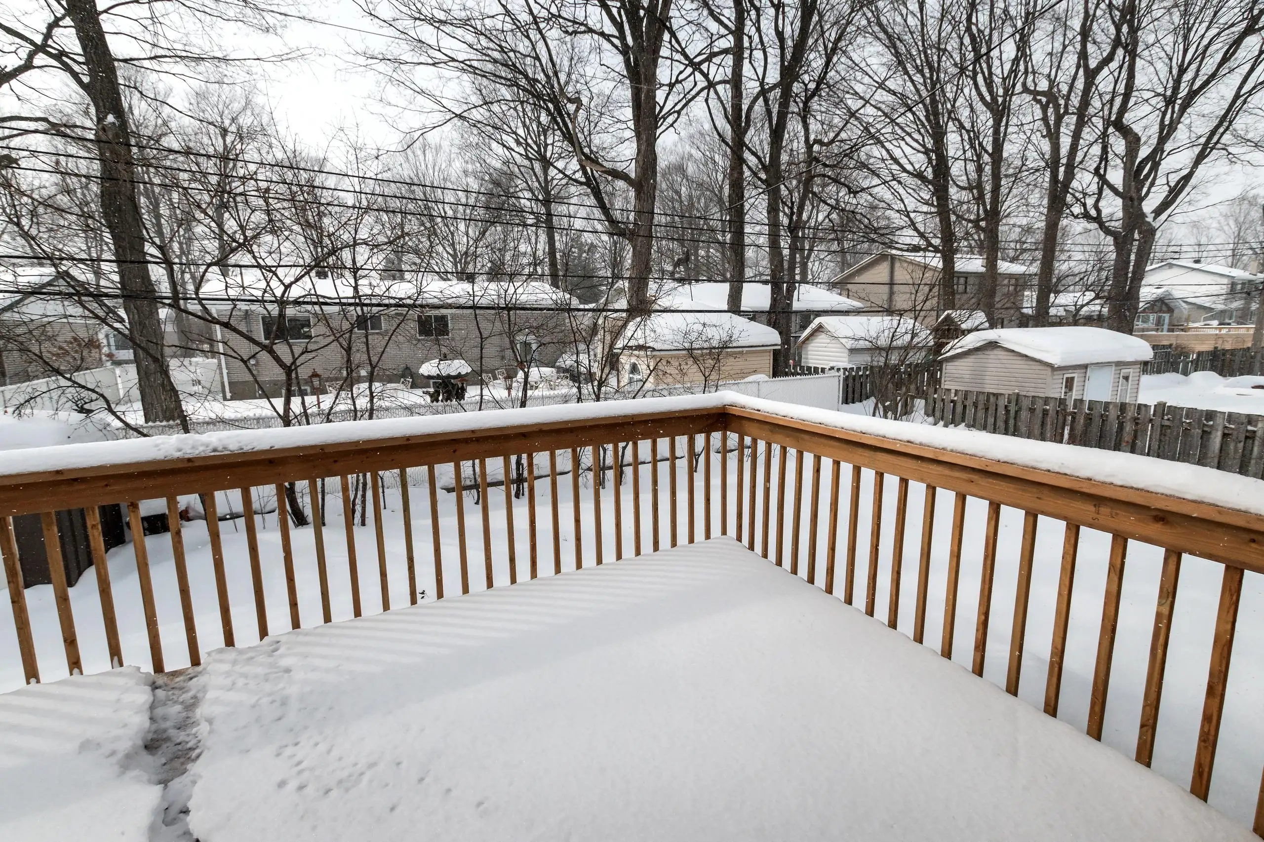 Best Time of Year to Build a Deck A deck covered in a thick blanket of snow. The view is looking out from the deck towards a residential neighborhood. In the background, there are houses and sheds also covered in snow, and numerous bare-branched trees.