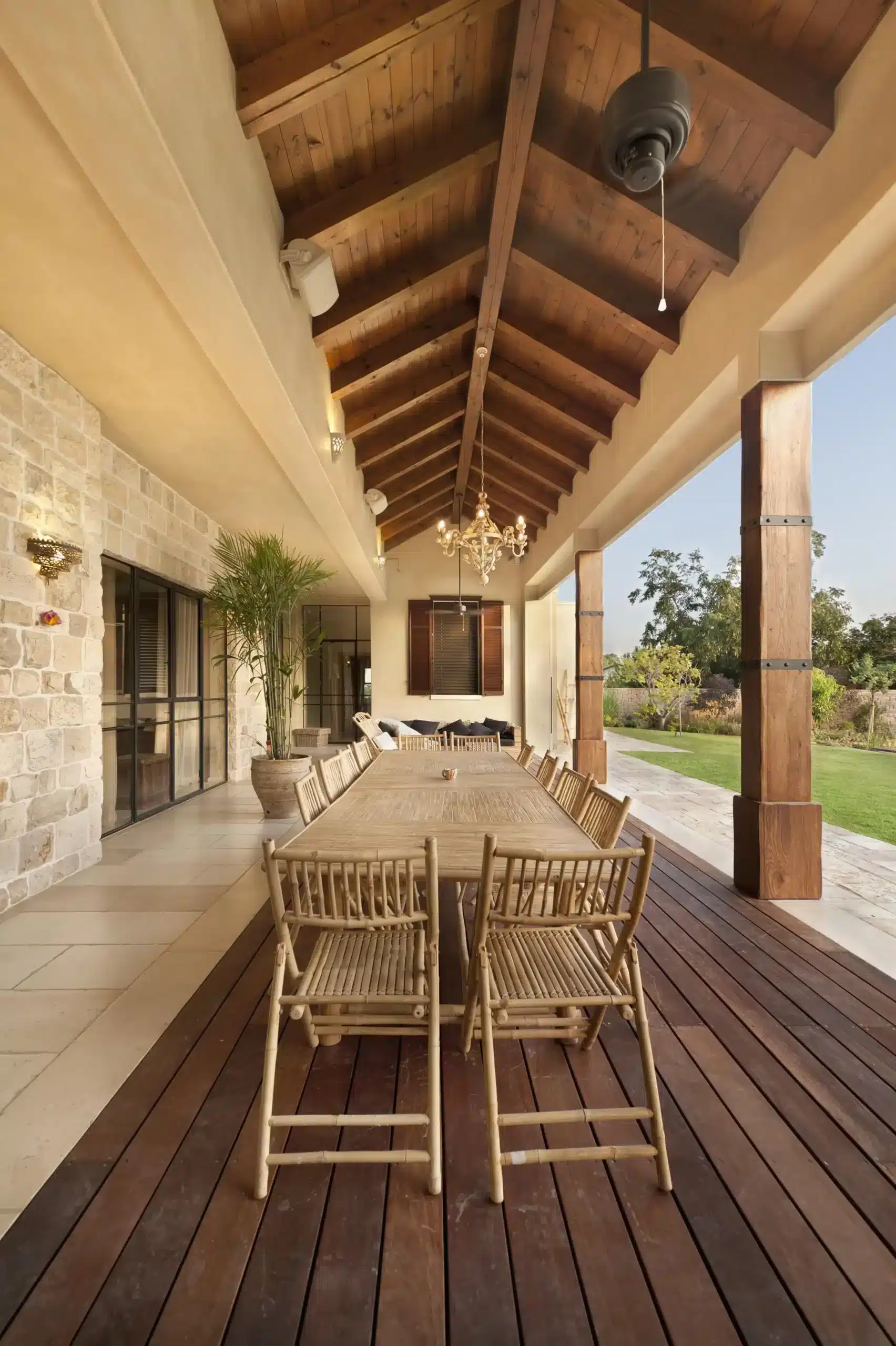 A long, covered outdoor veranda with a wide wood plank floor, supported by large wooden posts, featuring a lengthy bamboo dining table and folding chairs, with a stone facade on the house wall. The space is illuminated by a small chandelier and sunlight on the far end.