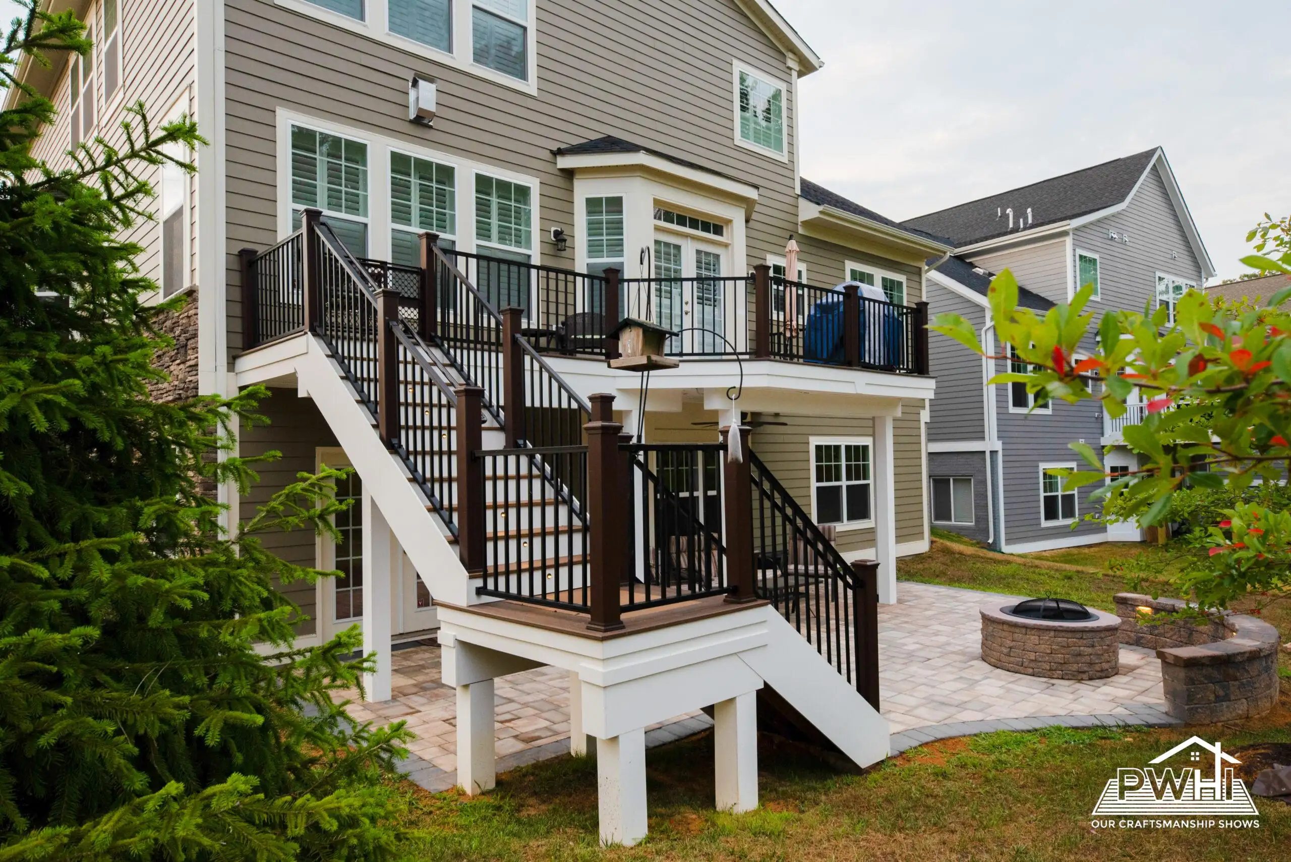 The exterior of a two-story house with a prominent multi-level deck and a ground-level patio. The house has light-colored siding, dark window frames, and several windows, including a bay window on the upper level. A set of stairs with dark railings leads from the upper deck down to a landing, and then another set of stairs descends to the ground.