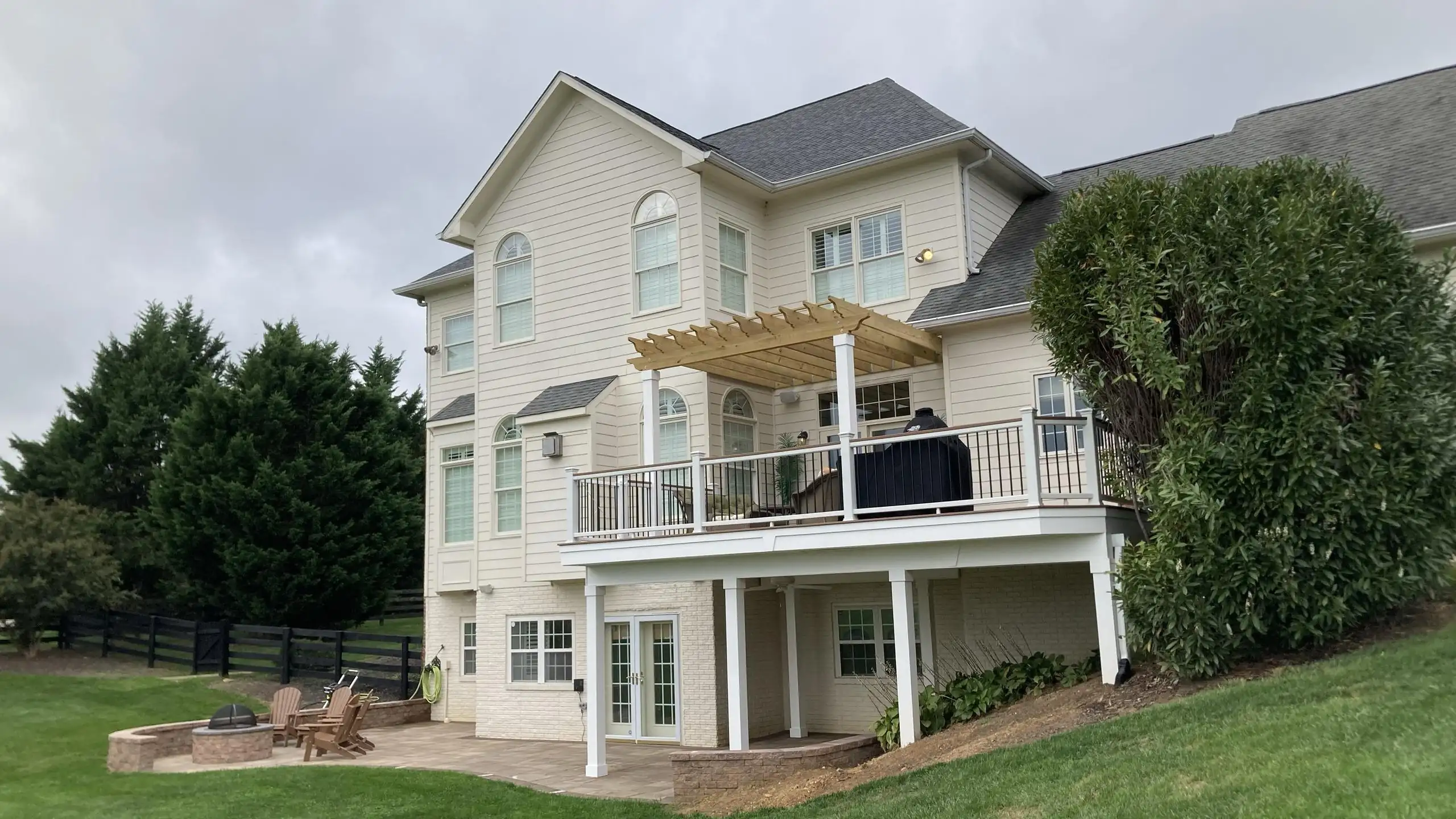 A large, light-colored house with an elevated deck and a pergola. The deck features white railings with black balusters and a dark-colored grill. A wooden pergola is built over a portion of the deck. Below the elevated deck, there's a patio area with a stone fire pit surrounded by wooden Adirondack chairs.
